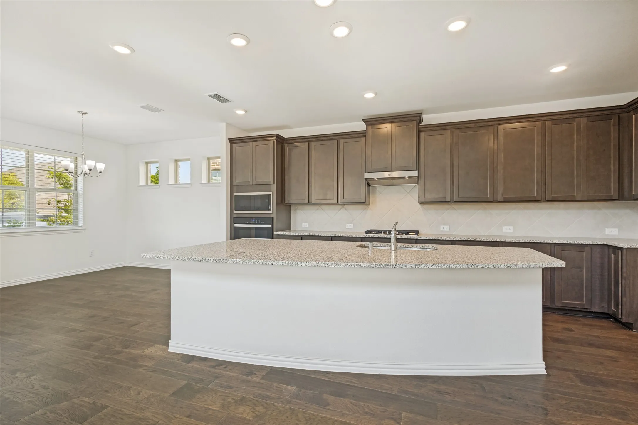 Kitchen featuring a large island with a light-toned countertop, dark wood-finish cabinetry, built-in double ovens, and a tile backsplash