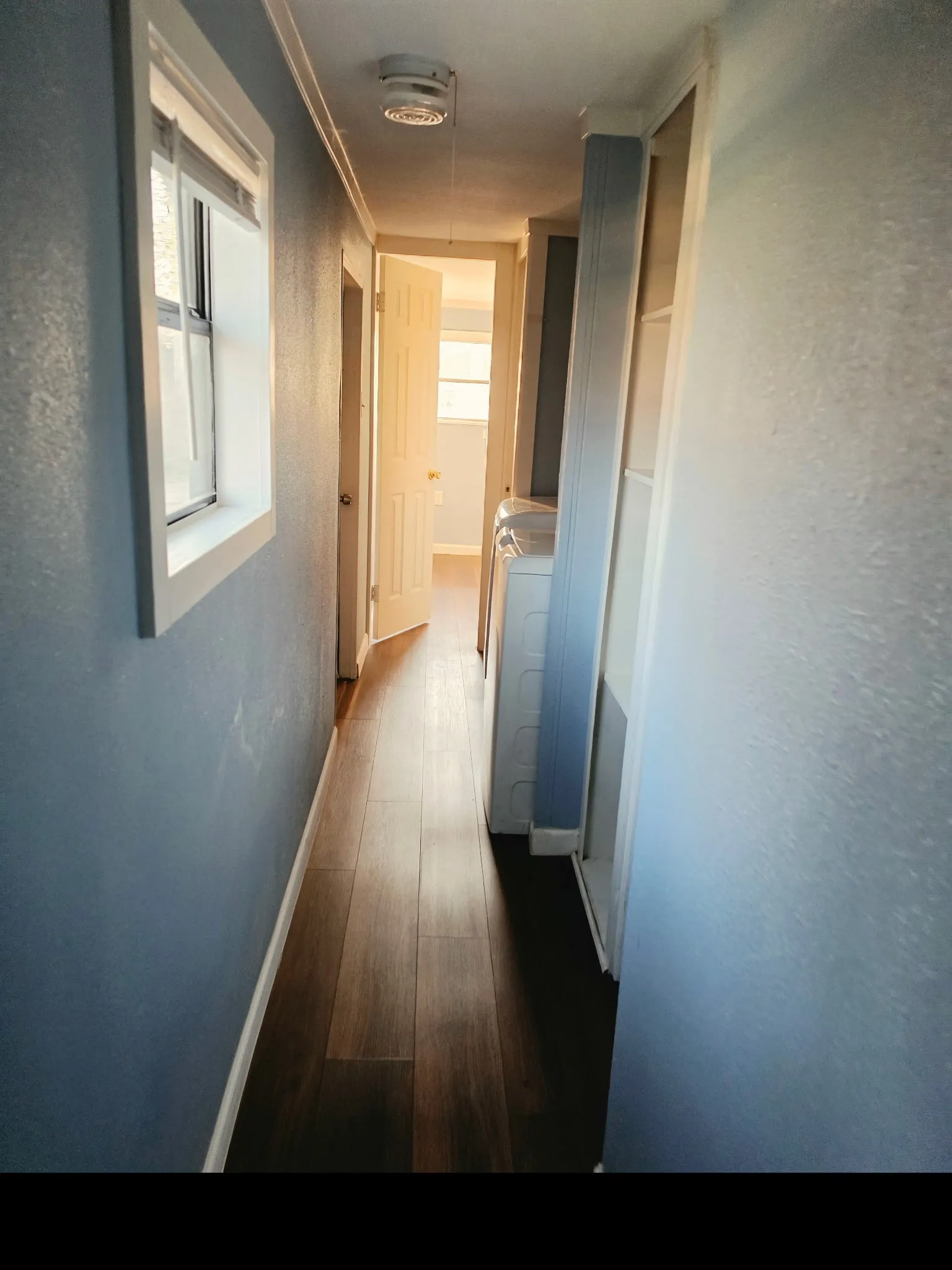 Hallway featuring wood-finish flooring, light blue wall paint, a window with white trim, and a built-in laundry area