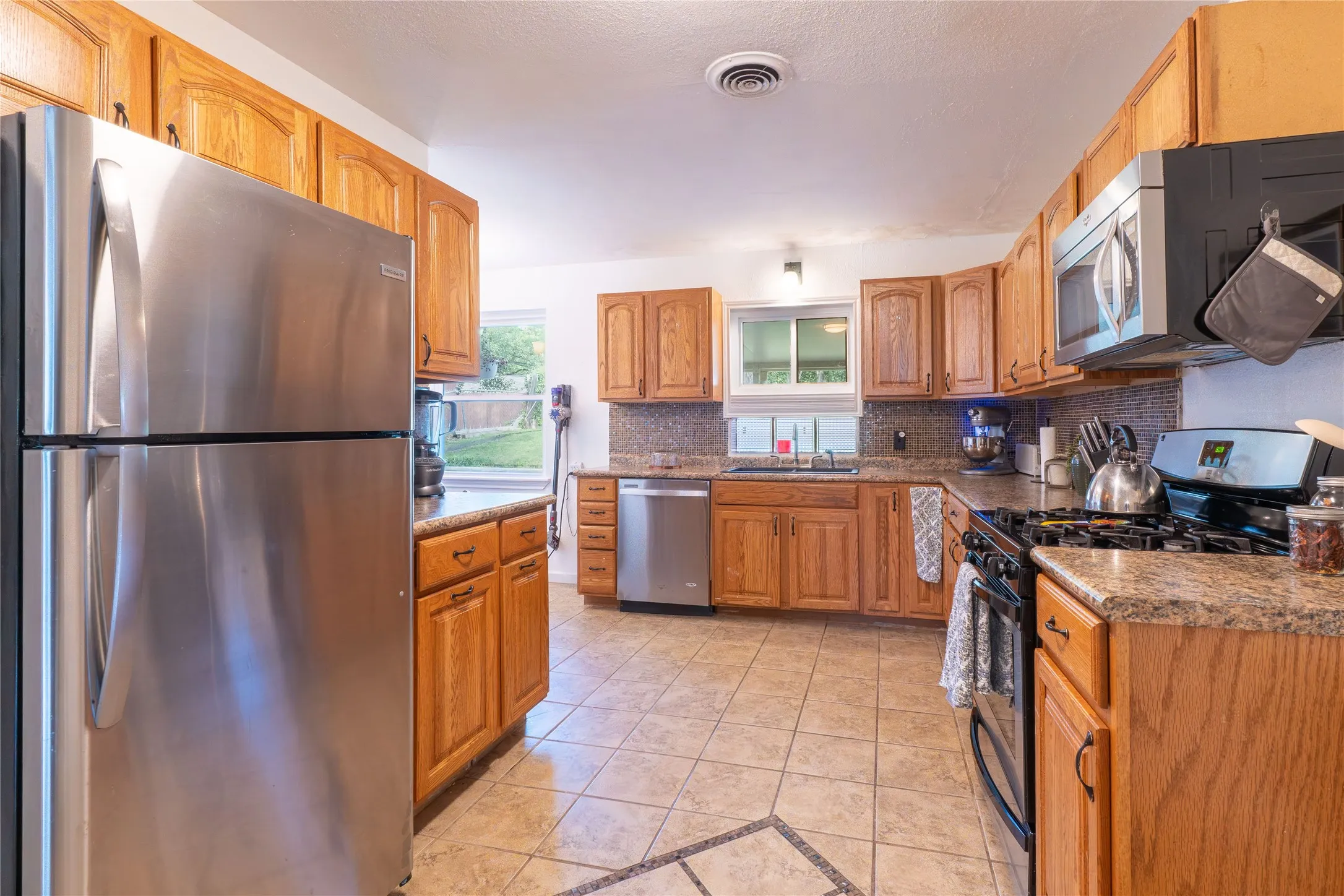 Kitchen featuring stainless steel appliances, light tile patterned floors, decorative backsplash, wood finish cabinetry, and dark stone countertops