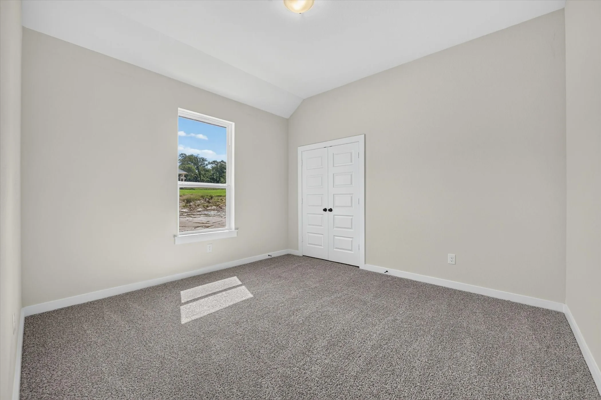 Spacious room featuring vaulted ceilings, a large window, neutral carpeting, light gray walls, and white bi-fold doors