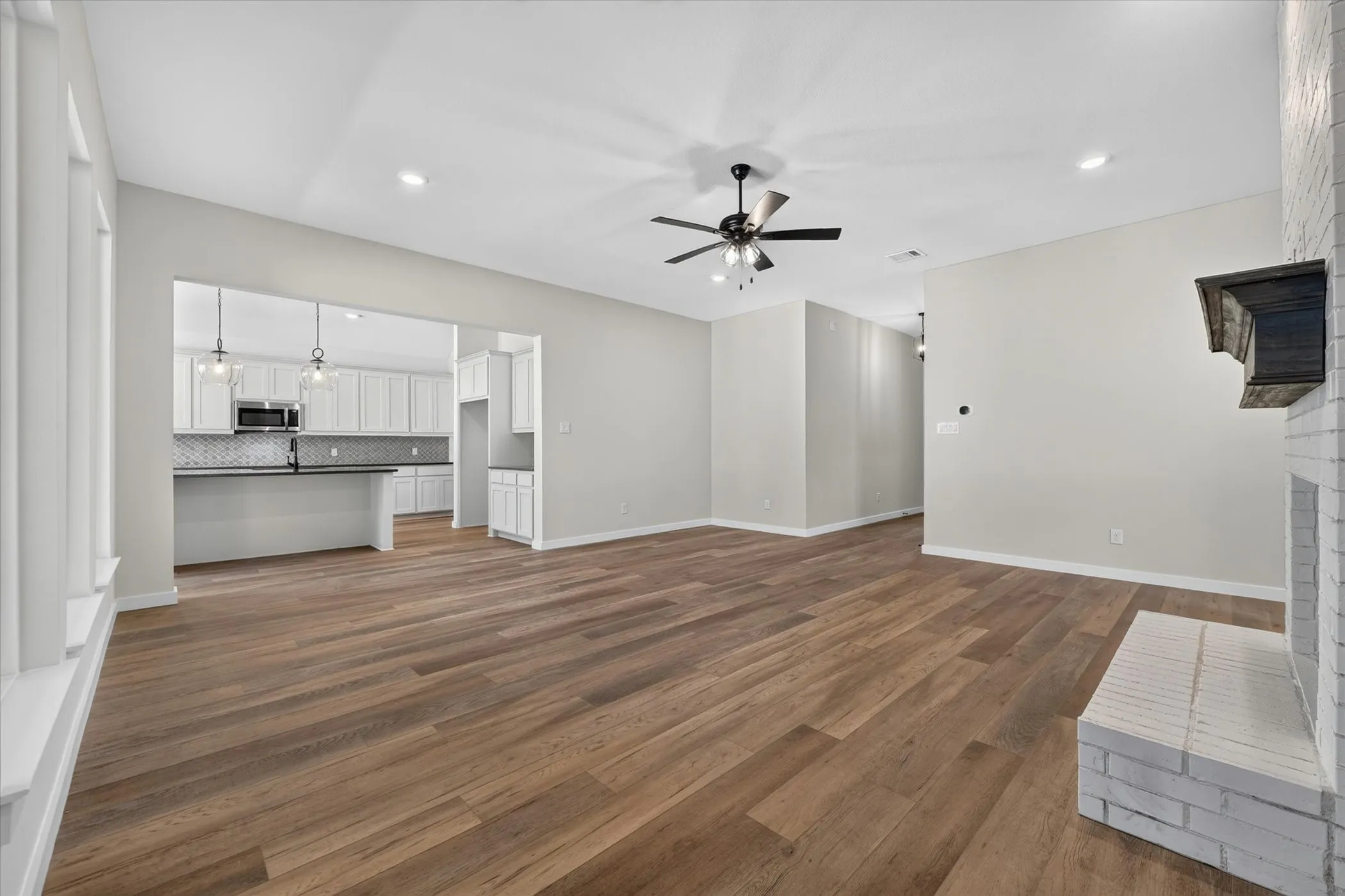 Spacious living area featuring wood-finish flooring, a white brick fireplace with a dark wood mantel, and an open concept layout to the kitchen