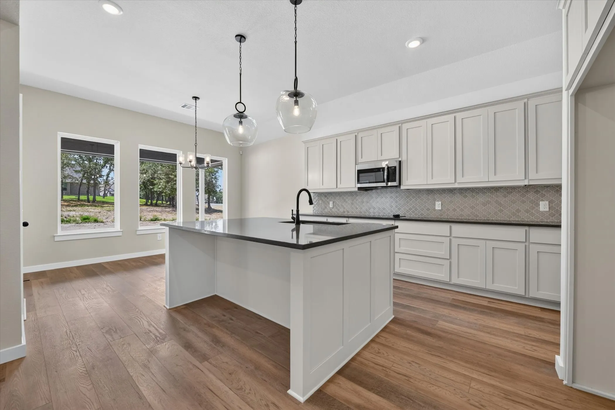 Spacious kitchen featuring a large central island with a sink, extensive white cabinetry, dark countertops, a tiled backsplash, and wood-finish flooring