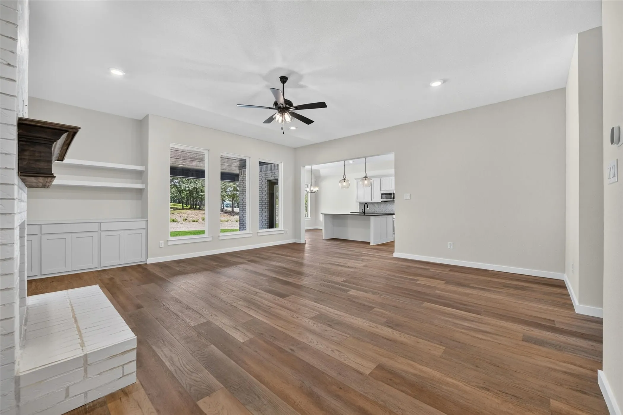Spacious living area featuring wood-finish flooring, a white brick fireplace with a dark wood mantle, and built-in cabinetry with open shelving