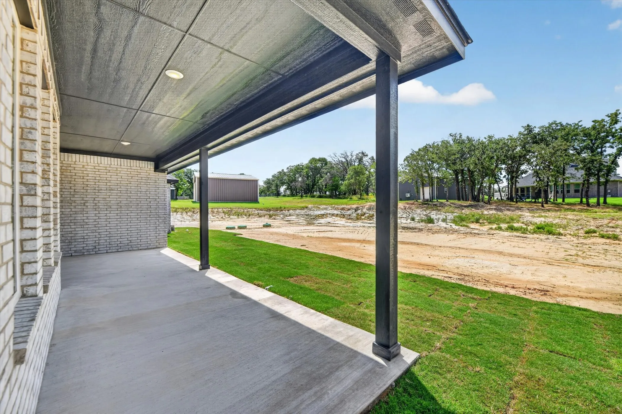 Covered concrete patio with recessed lighting, supported by dark posts, featuring a light brick exterior and an expanse of green lawn