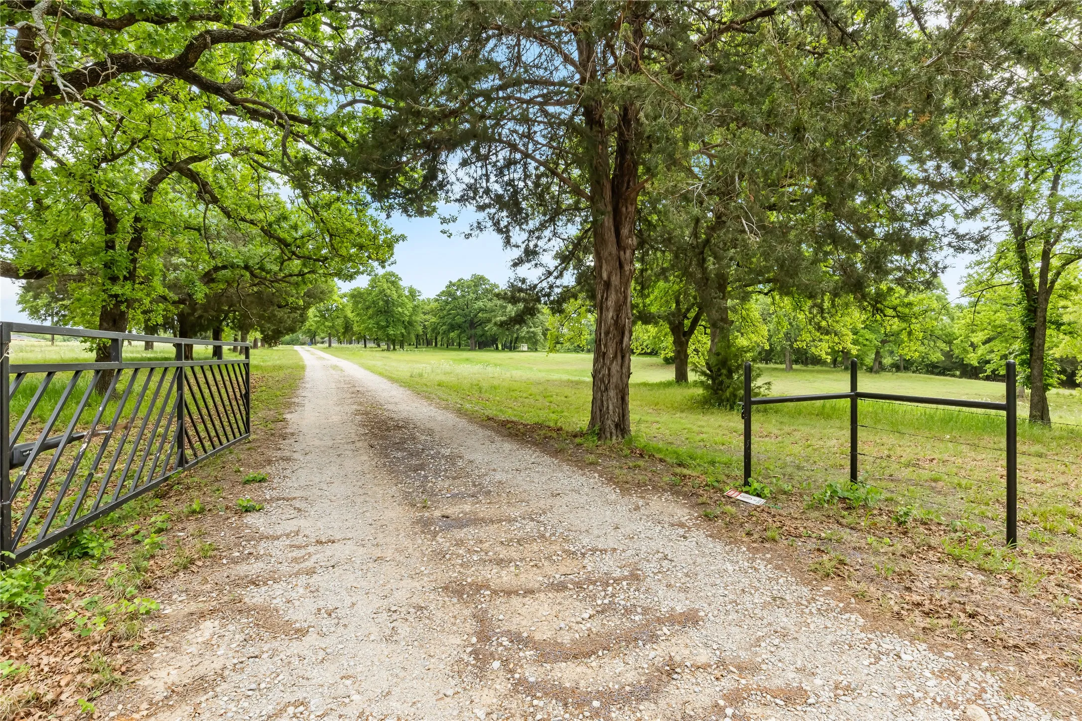 A gravel driveway leads through a gated entrance, bordered by a green lawn and mature trees