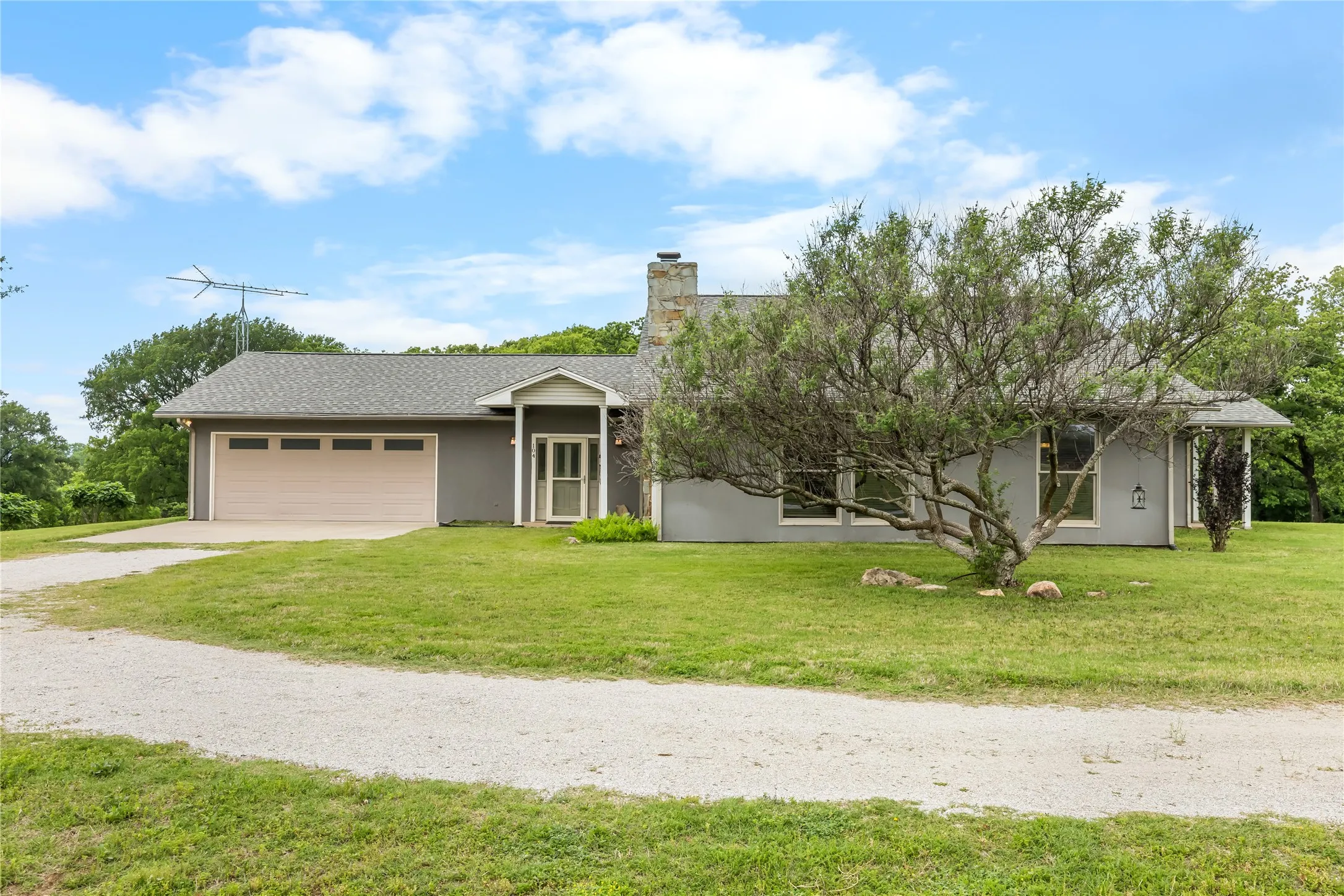 The property features a grey exterior with a tan garage door, a gravel driveway, and a well-maintained lawn