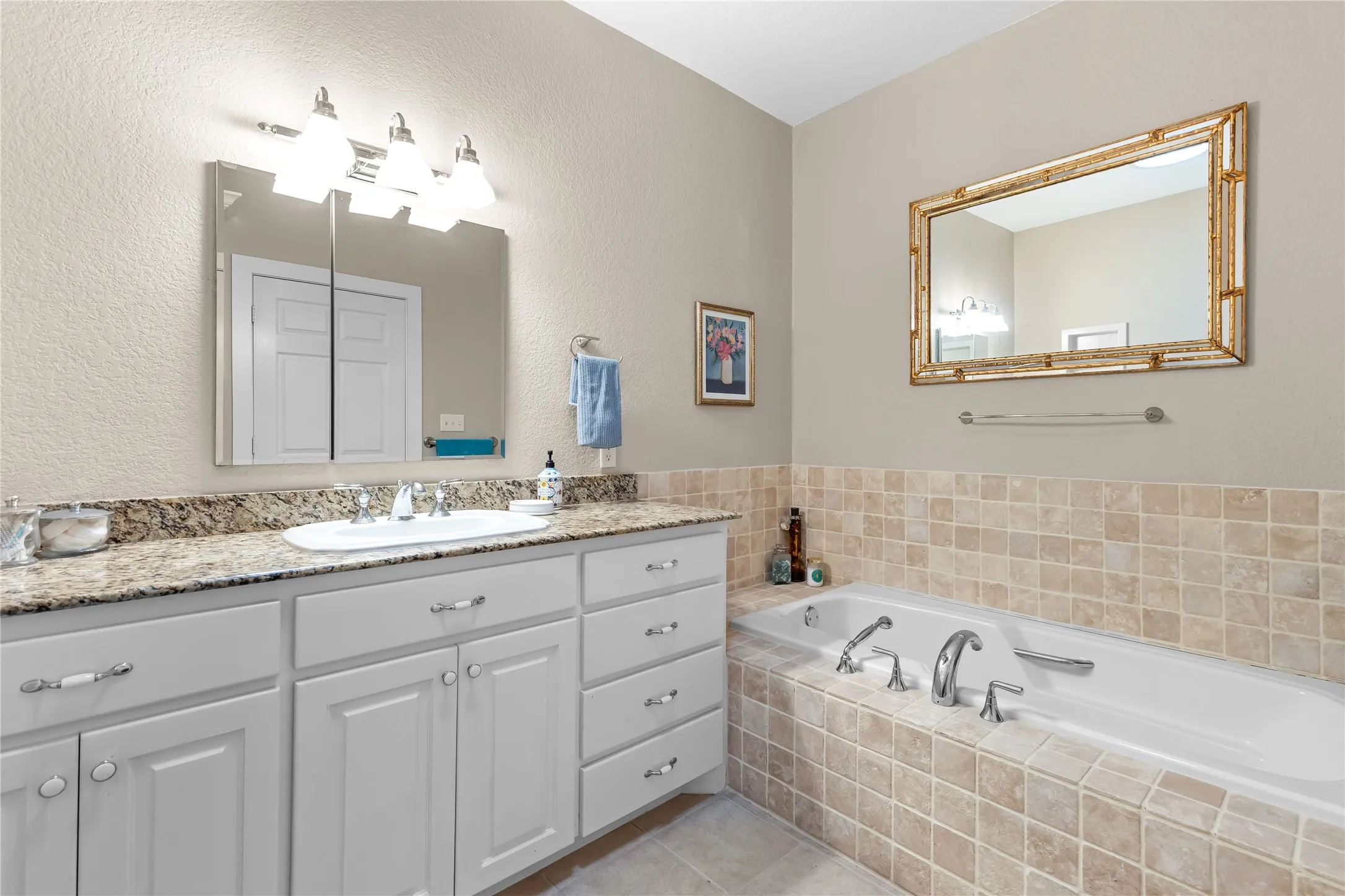 The bathroom features a spacious vanity with white cabinetry and a granite countertop, a bathtub surrounded by square tiles, and a framed mirror above the tub