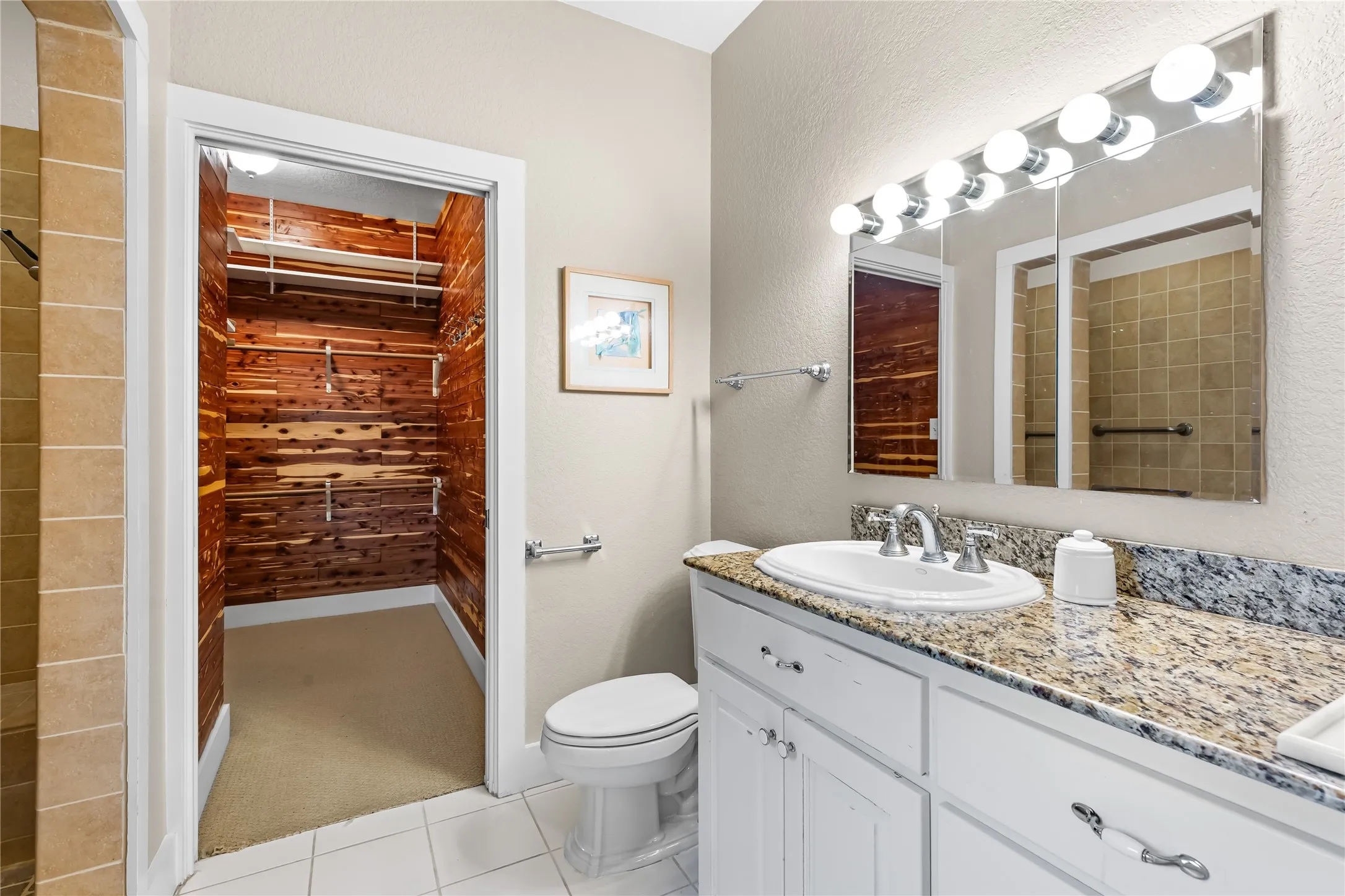 The bathroom features a vanity with a granite countertop and a built-in sink, a large mirror with overhead lighting, cedar closet and white tile flooring