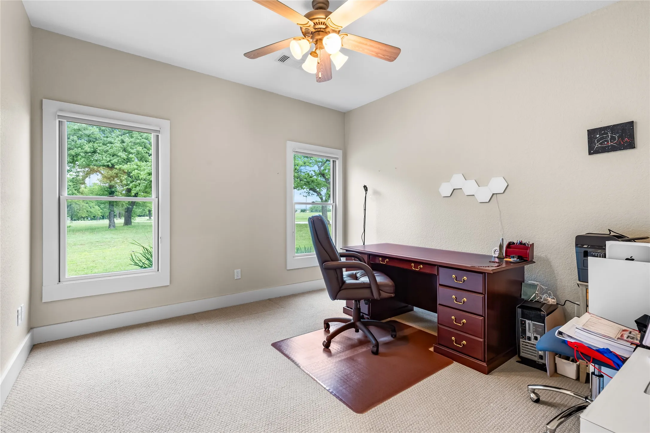 This room features two windows providing views of the surrounding greenery, a ceiling fan, and light-colored carpeting