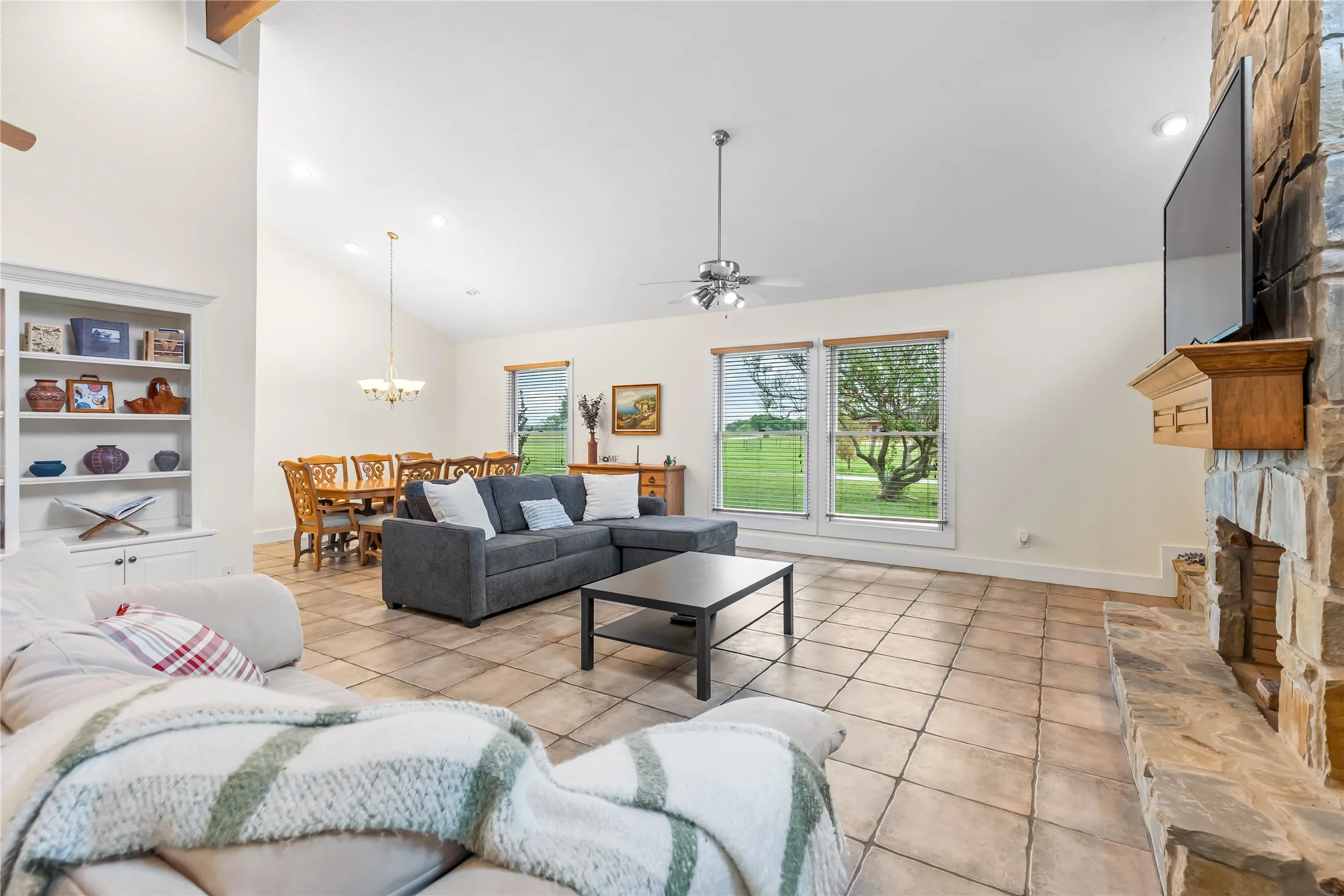 Spacious living area featuring vaulted ceilings, tiled flooring, a stone fireplace, and large windows providing views of the green landscape