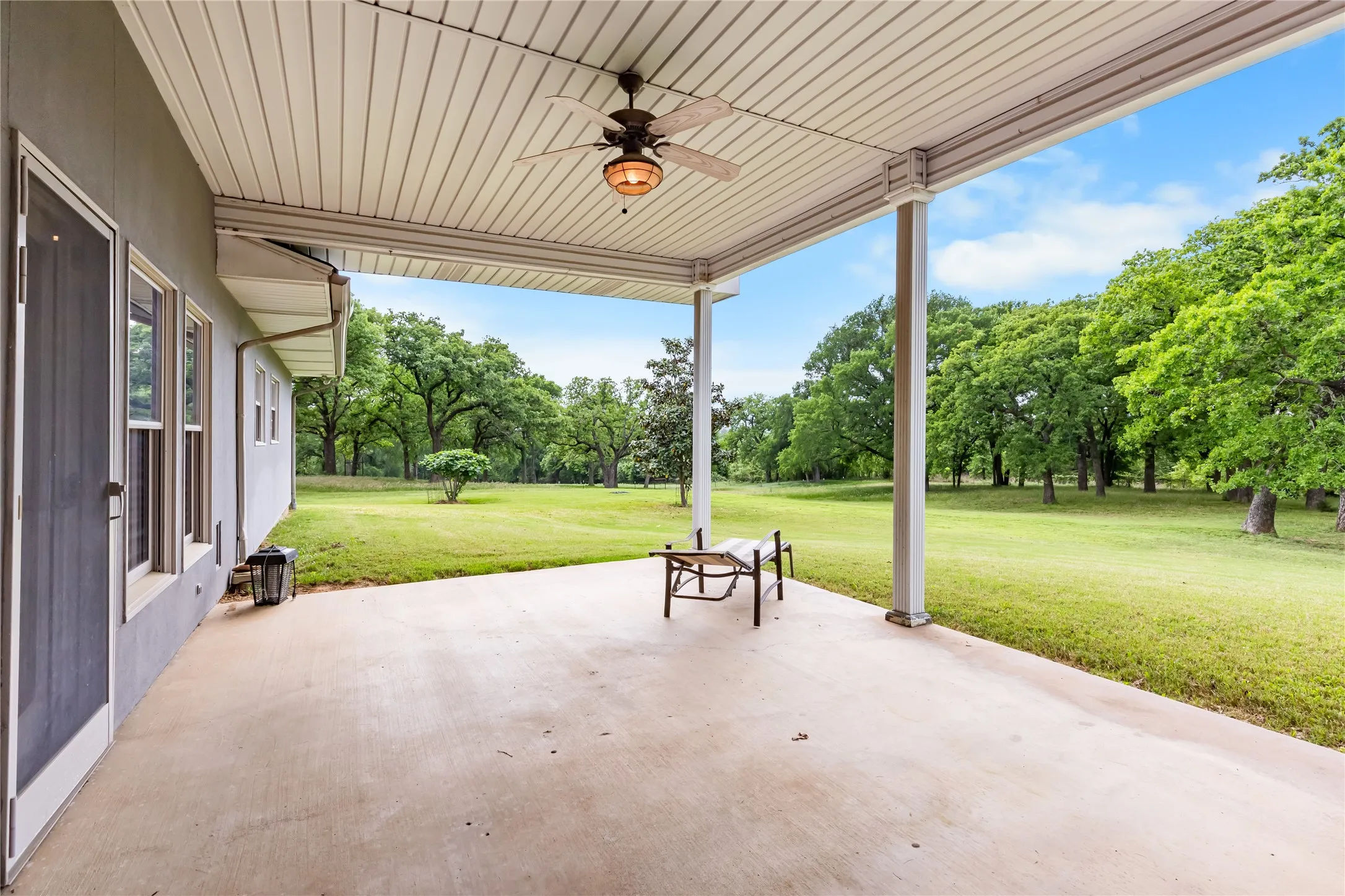 This property features a covered patio with a ceiling fan, overlooking an expansive grassy lawn and mature trees