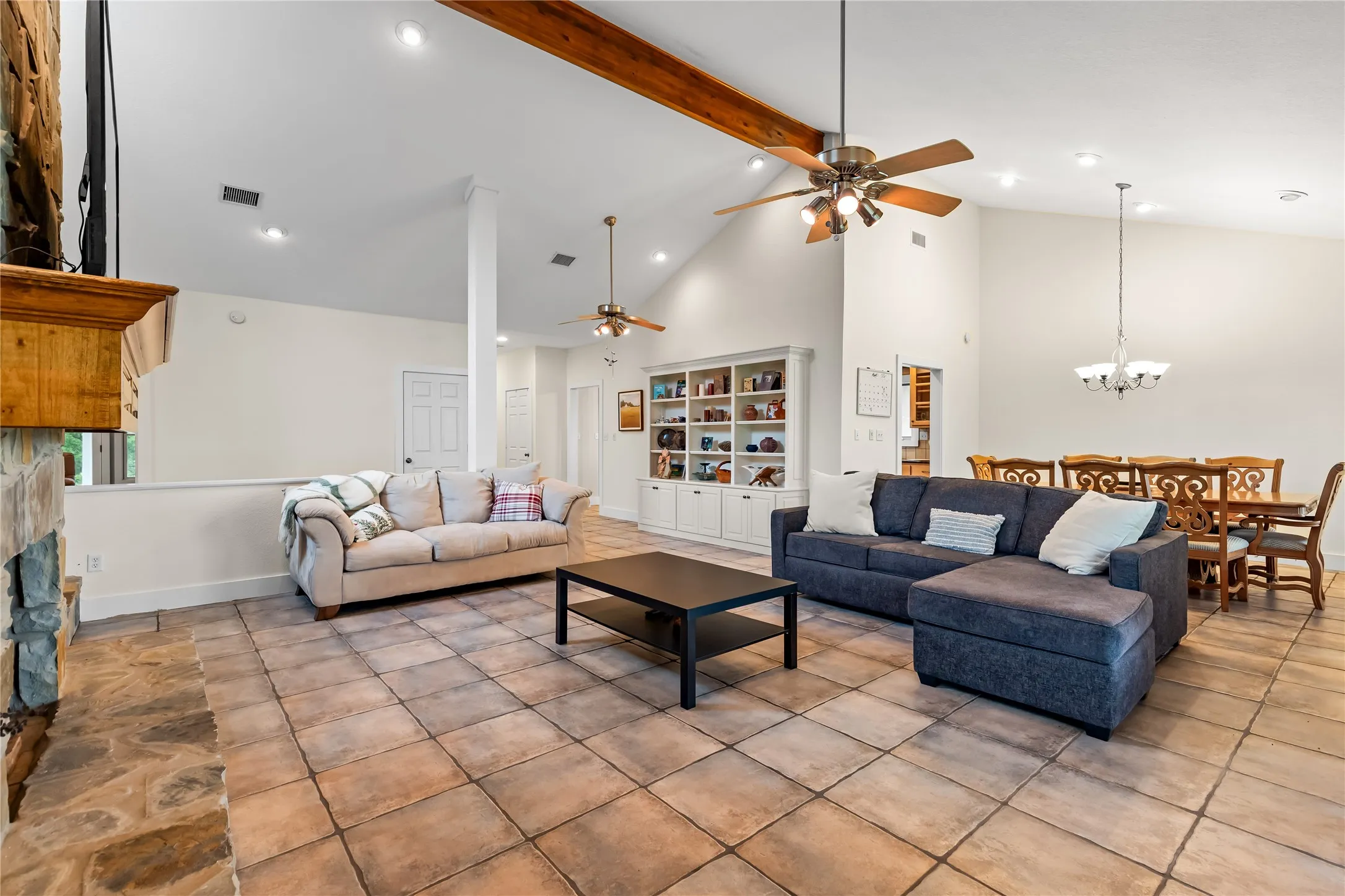 Spacious living area featuring tiled flooring, a stone fireplace, vaulted ceilings with a wood beam, and recessed lighting