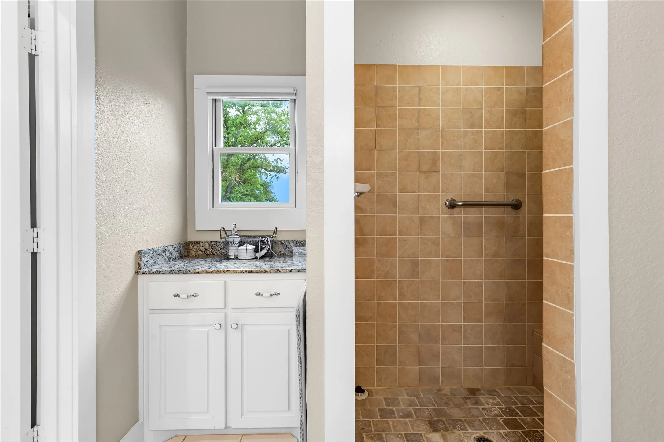 The bathroom features a vanity with a granite countertop and a window providing natural light, alongside a tiled shower with a grab bar and a tiled floor