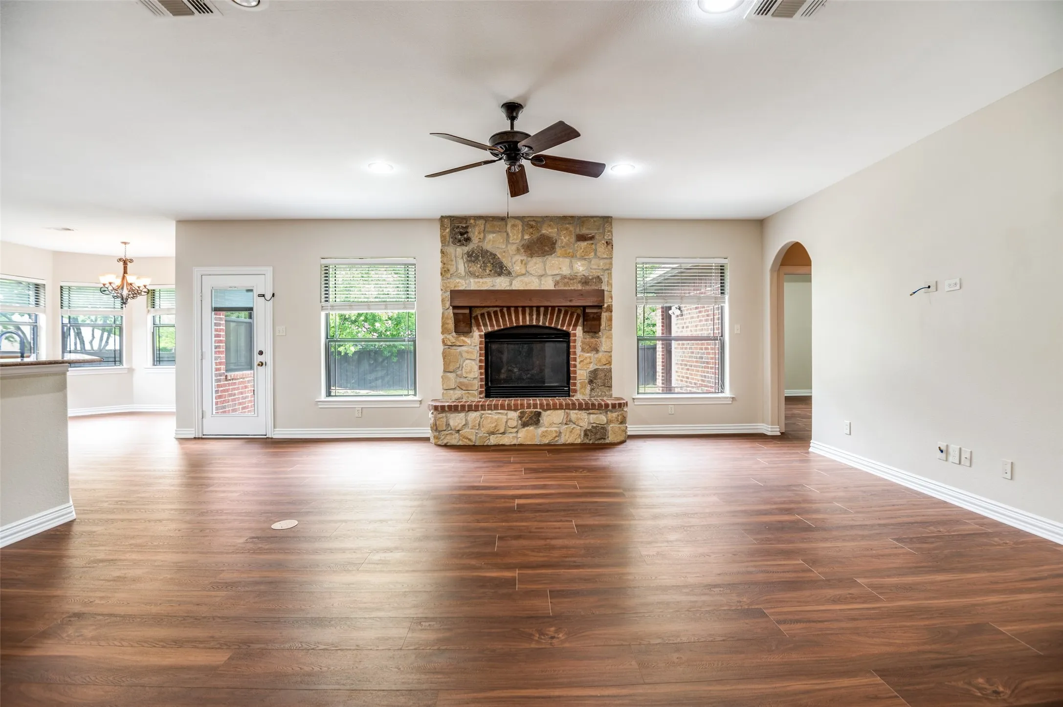 Spacious living area featuring wood-finish flooring, a stone fireplace with a substantial wood mantel, a ceiling fan, and multiple windows