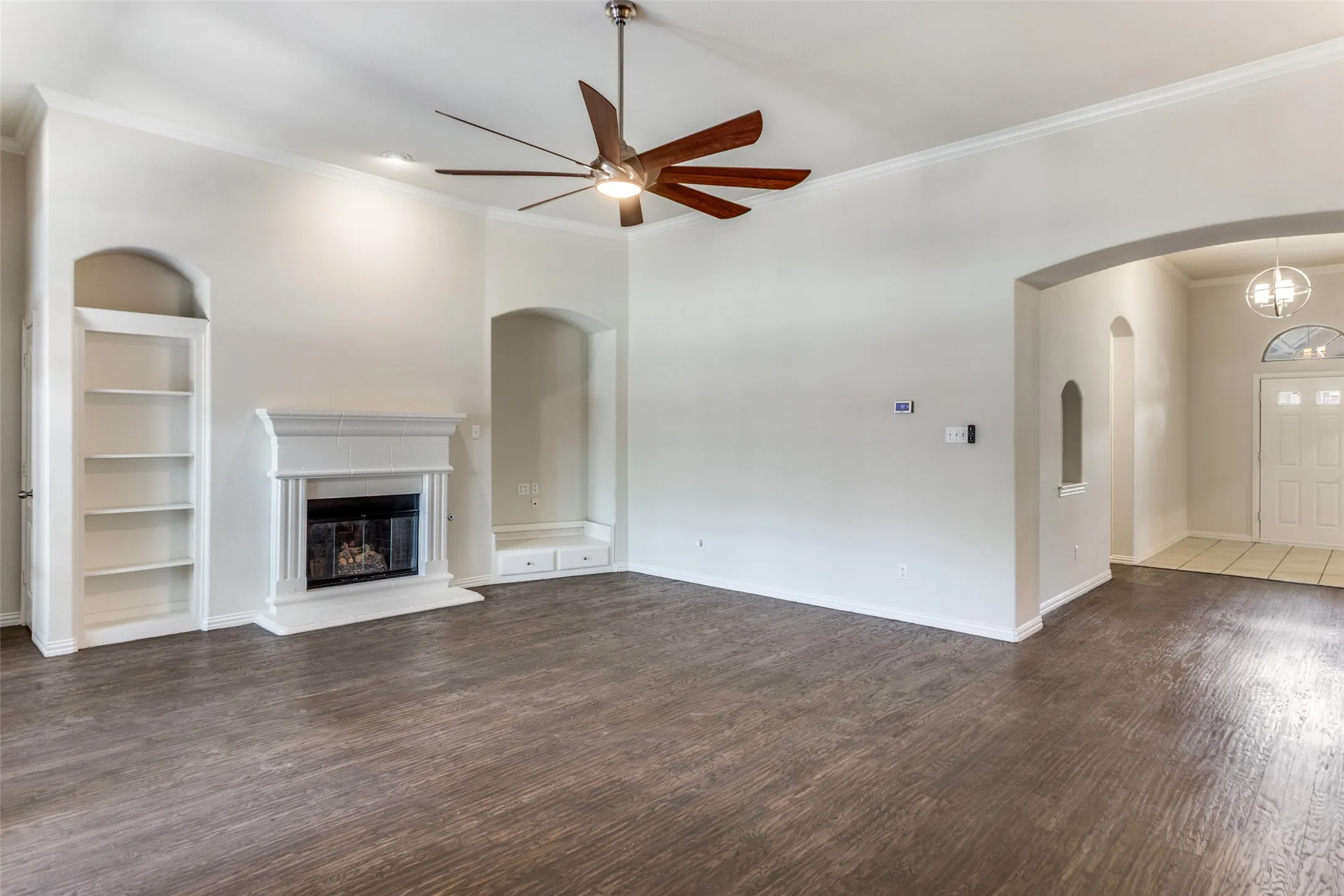 living room with built in shelves, crown molding, dark wood-style floors, ceiling fan, and a glass covered fireplace