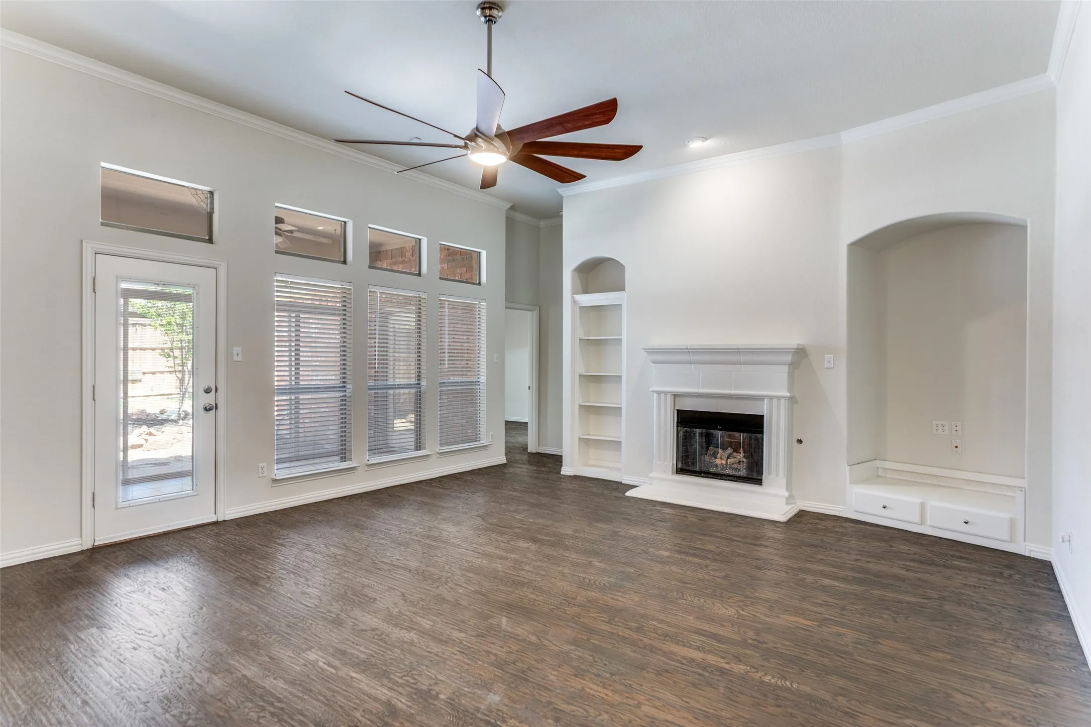 living room featuring built in shelves, ceiling fan, a glass covered fireplace, crown molding, and dark wood finished floors