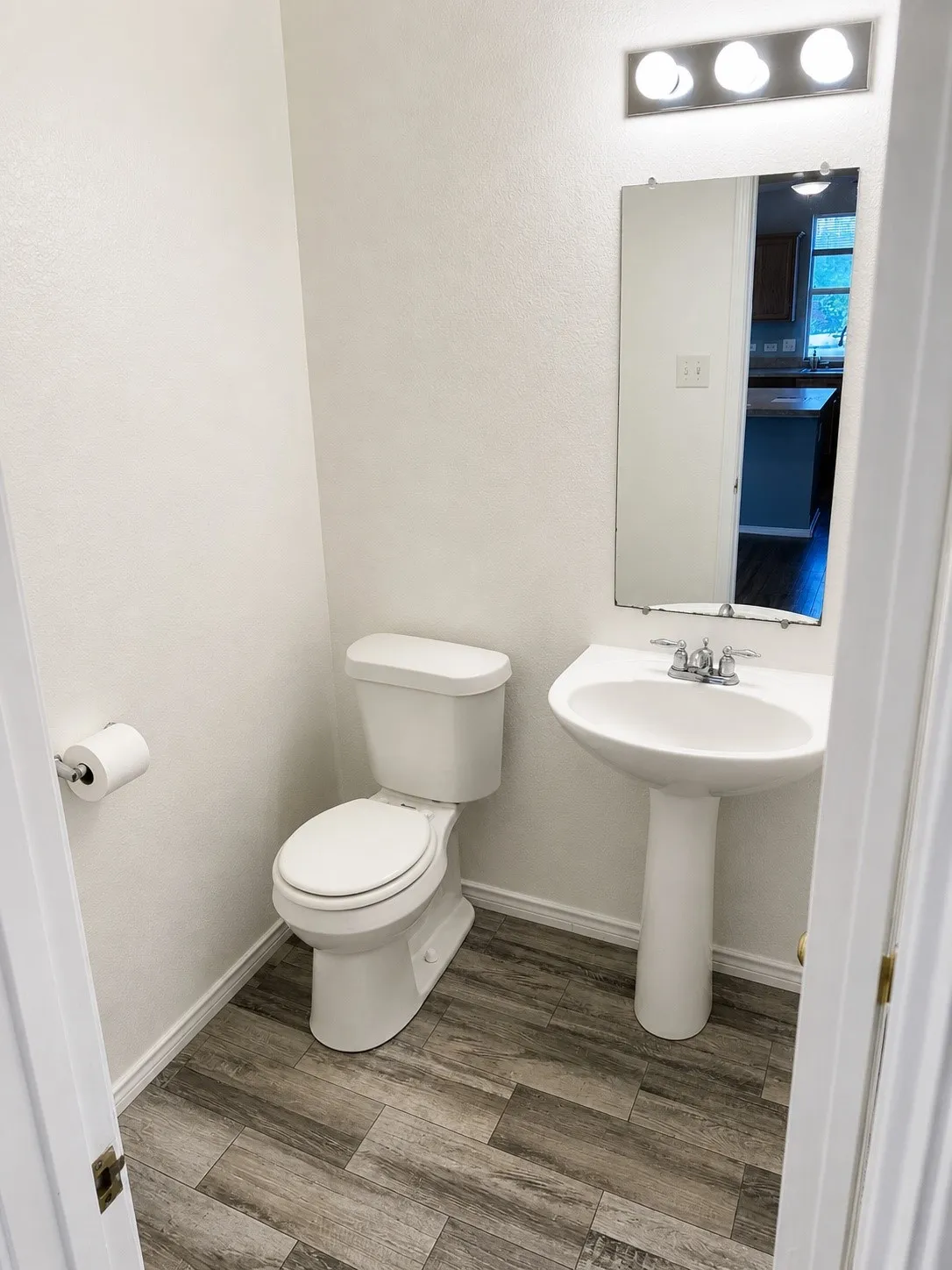 Powder room featuring a pedestal sink, white toilet, wall-mounted mirror, and wood-finish flooring
