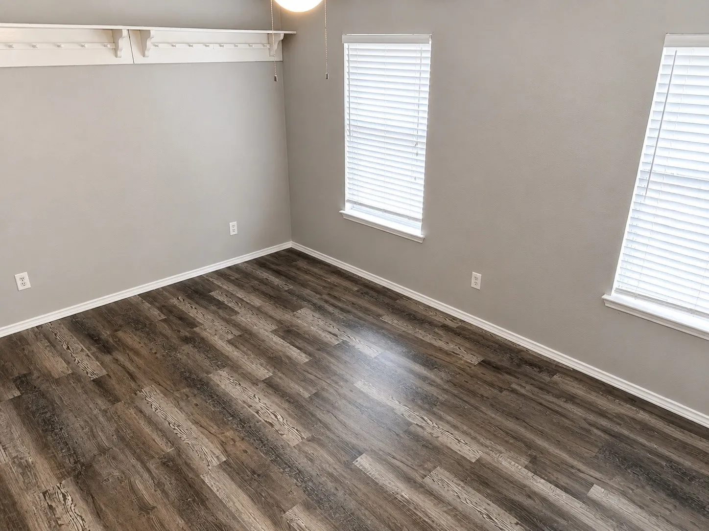 Room featuring wood-finish flooring, light grey walls, white baseboards, and two windows with blinds