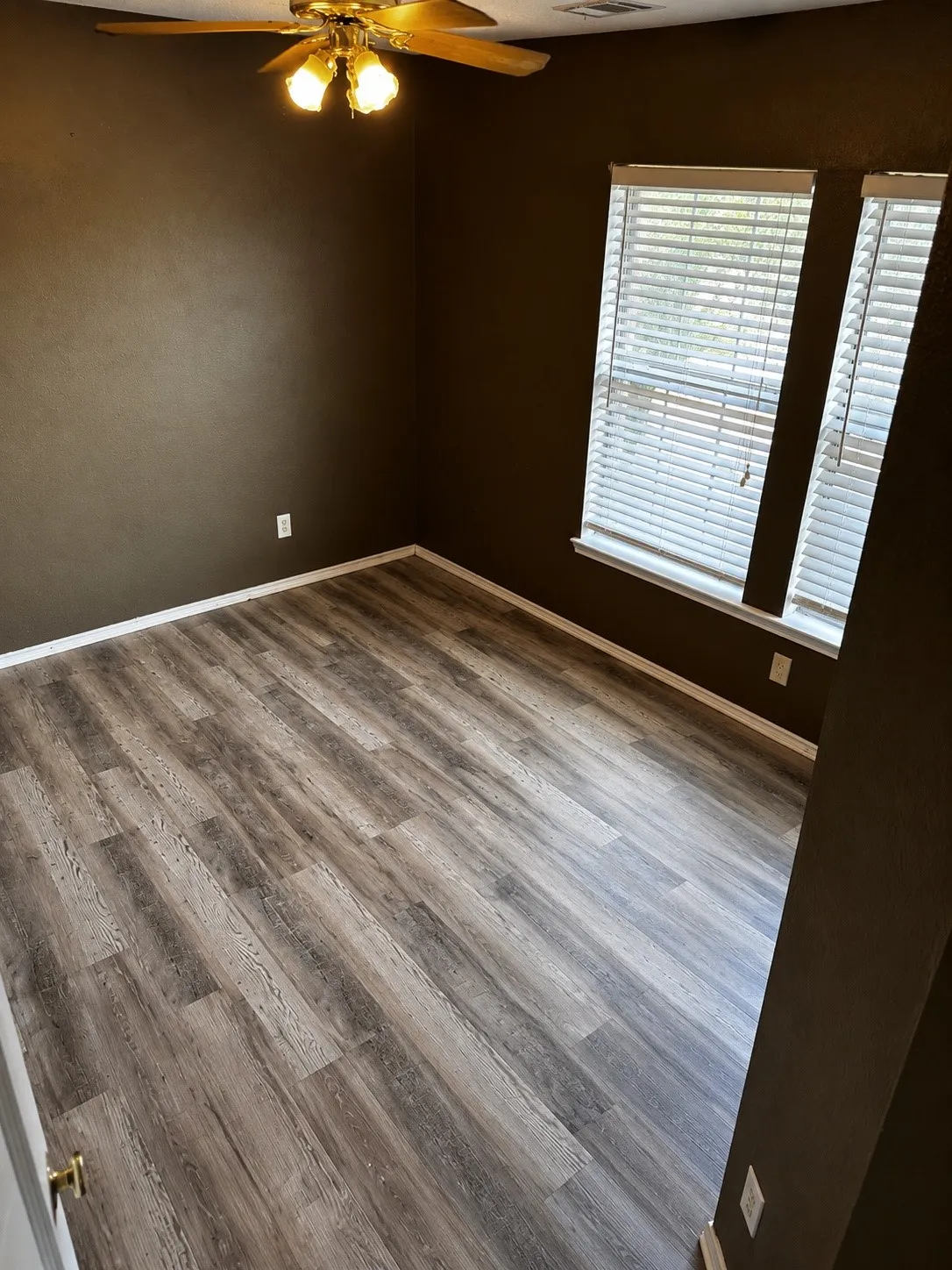 Room featuring wood-finish flooring, two large windows with blinds, a ceiling fan with integrated lighting, and dark-toned walls