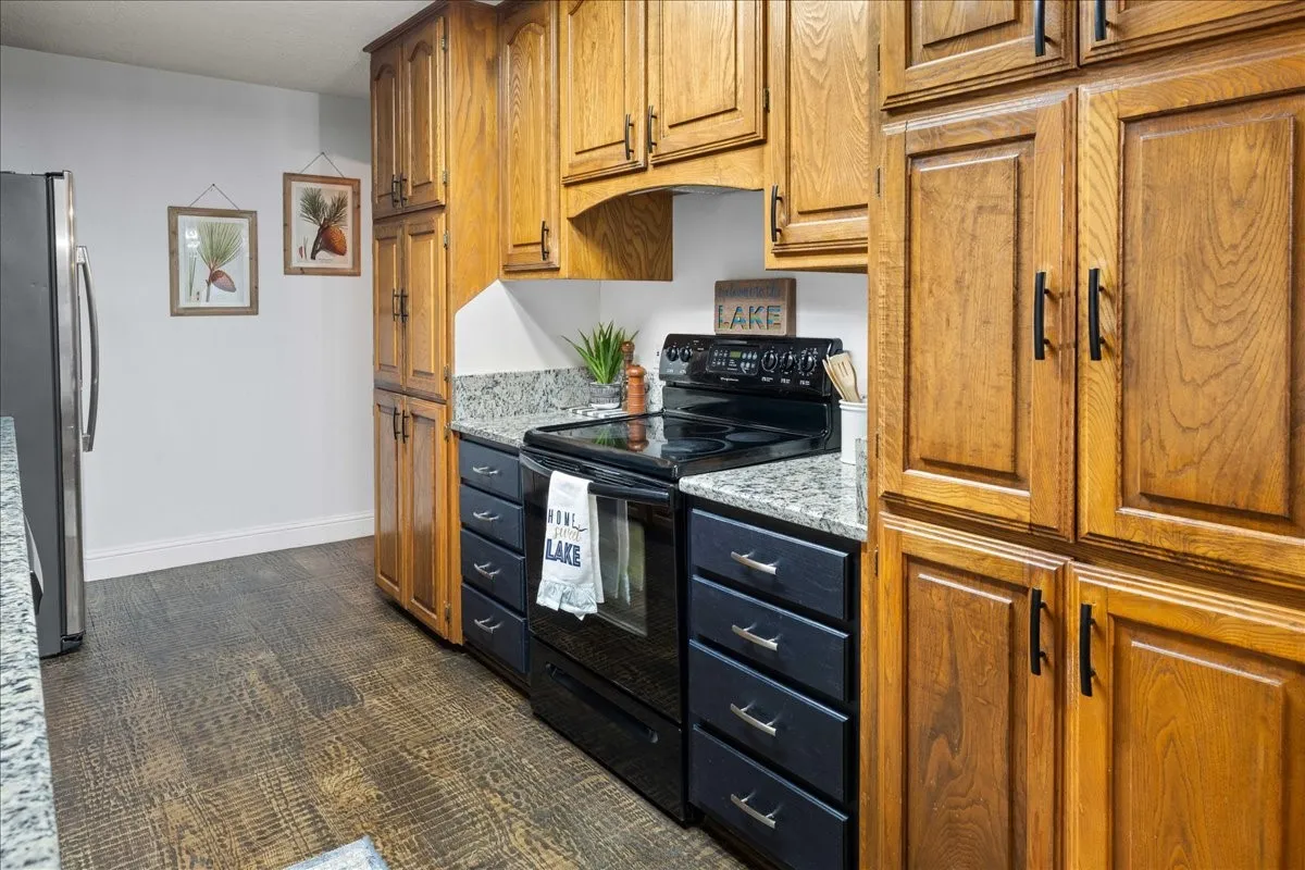 Kitchen with black electric range oven, brown cabinetry, light stone counters, freestanding refrigerator, and dark carpet