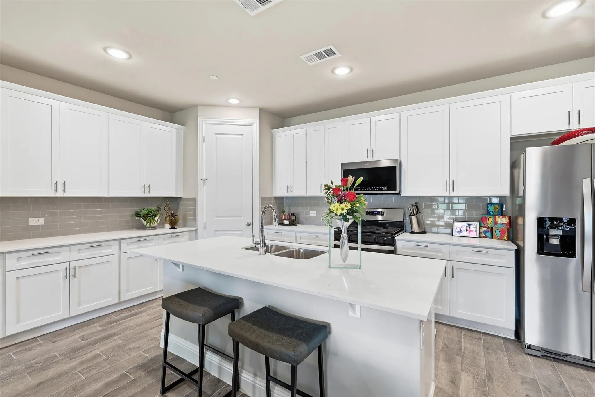 . The Cook’s Kitchen showcases Modern White Shaker-Style Cabinetry, Light-Toned Quartz Countertops, a Spacious Center Island with Breakfast Bar, Soft Gray Subway Tile Backsplash, Recessed Lighting, and Upgraded Wood-Look Tile Flooring for Durability and Warmth. A Full-Height Pantry and Multiple Work Zones enhance Functionality and Flow, seamlessly connecting the Living and Dining Areas for Effortless Entertaining
