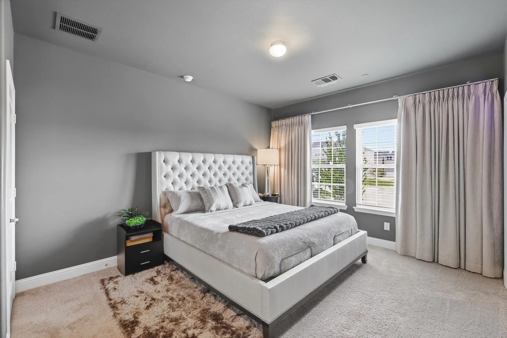 The Primary Bedroom offers a Spacious Layout, Soft Neutral Color Palette, Accent Wall Finish, Large Windows, and Full-Length Drapery, creating a Calm and Private Retreat with Natural Light and Modern Architectural Lines.