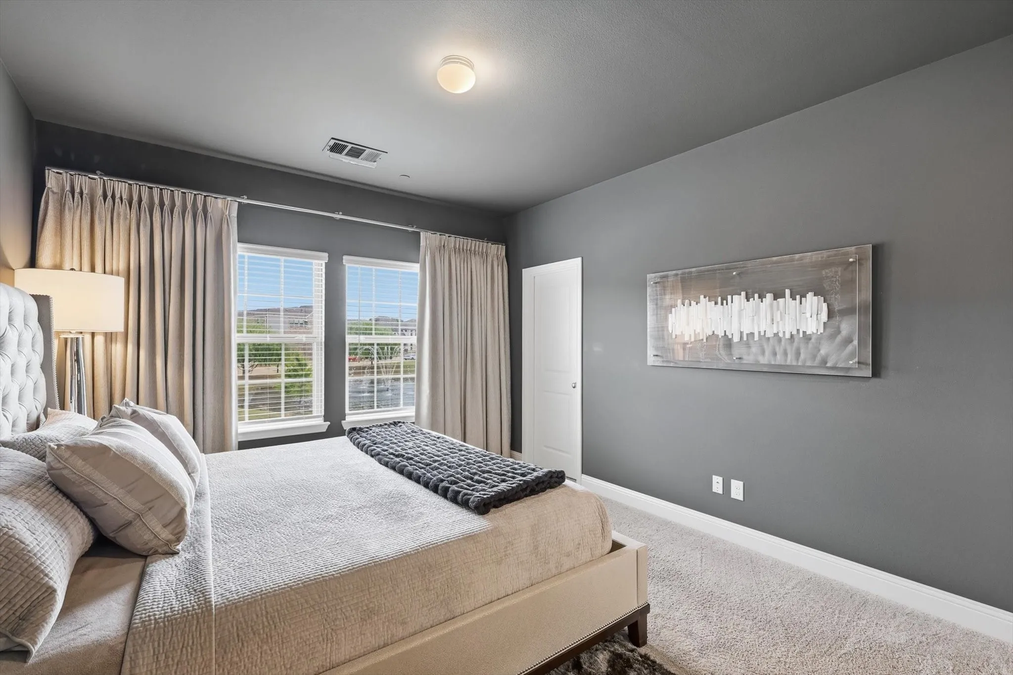The Primary Bedroom offers a Spacious Layout, Soft Neutral Color Palette, Accent Wall Finish, Large Windows, and Full-Length Drapery, creating a Calm and Private Retreat with Natural Light and Modern Architectural Lines.
