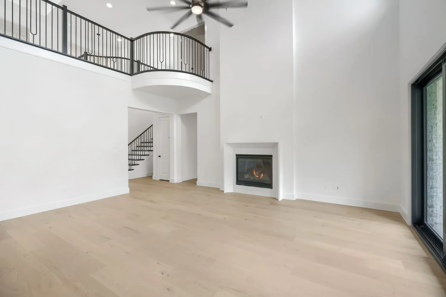 Light wood-finish flooring extending through a spacious room featuring a two-story ceiling, a contemporary fireplace, and a large window
