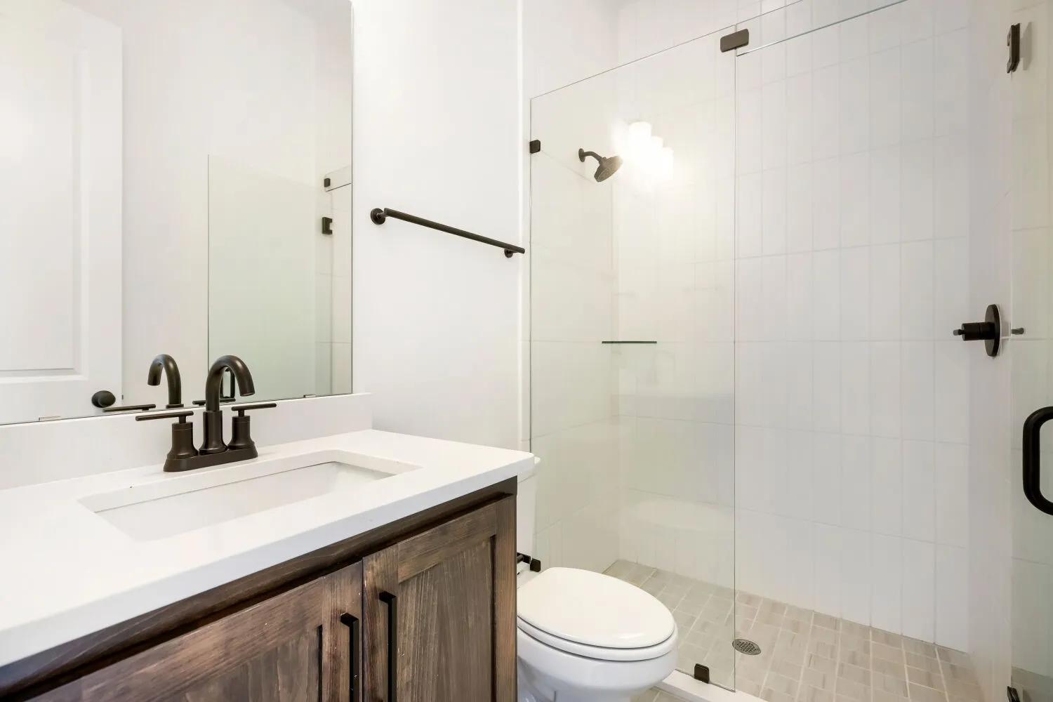 Bathroom featuring a dark wood-finish vanity with a white countertop and undermount sink, oil-rubbed bronze fixtures, a full-width mirror, and a glass-enclosed shower with white subway tile walls and a light-toned tile floor