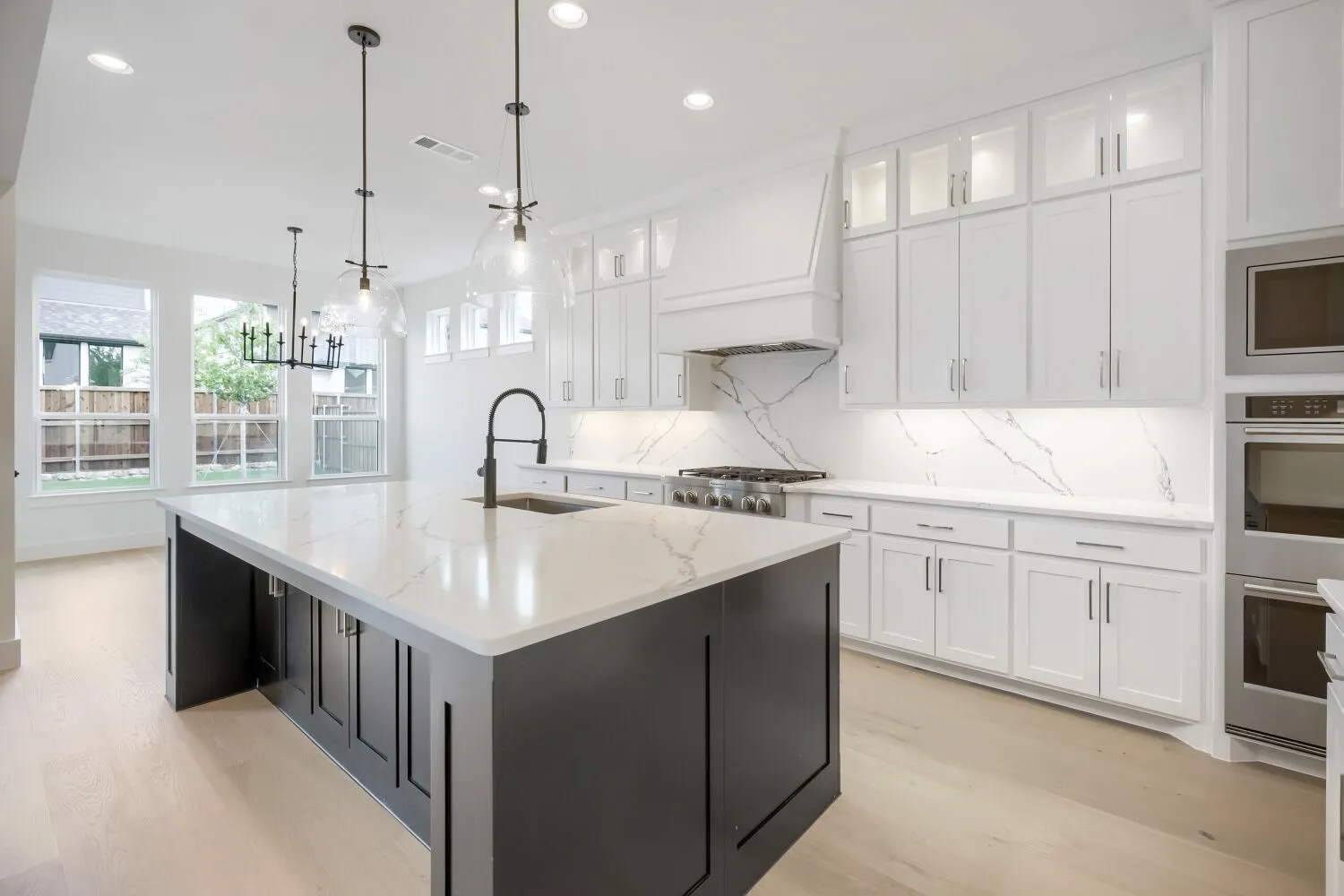 Gourmet kitchen featuring a large central island with a waterfall edge, stainless steel double ovens, a built-in gas range, white shaker cabinetry with upper glass-front cabinets, and light wood-finish flooring