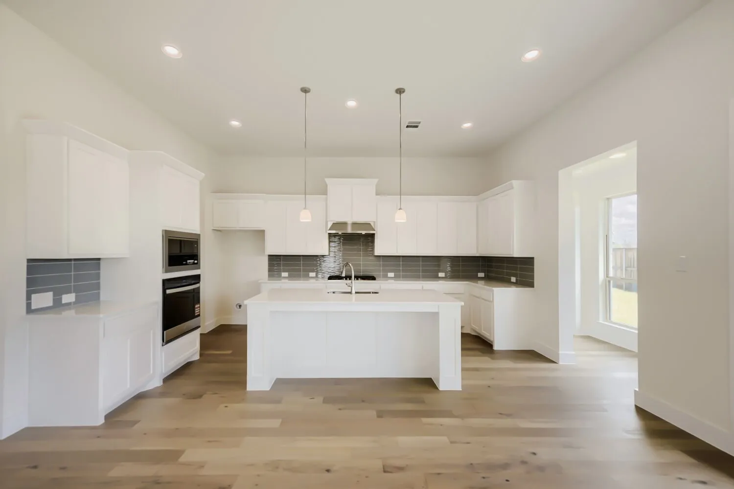 Contemporary kitchen featuring white cabinetry, a spacious island with sink, stainless steel appliances including a built-in oven and microwave, and a gray subway tile backsplash