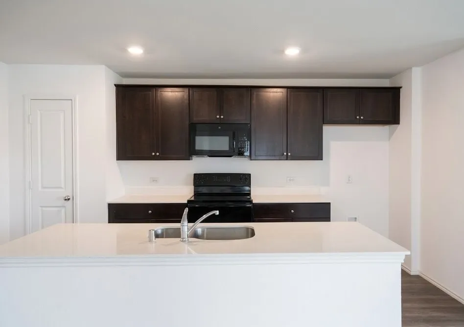 Kitchen with dark wood finish cabinets, black appliances, a center island with sink, light stone counters, and recessed lighting