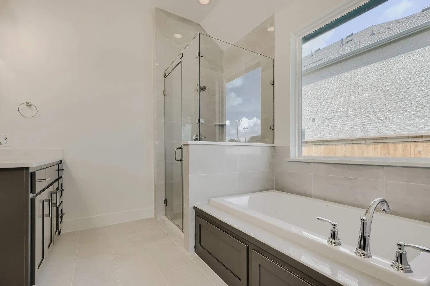 Bathroom featuring a built-in soaking tub with chrome fixtures, a glass-enclosed shower, a double vanity with dark cabinetry, and neutral tile flooring