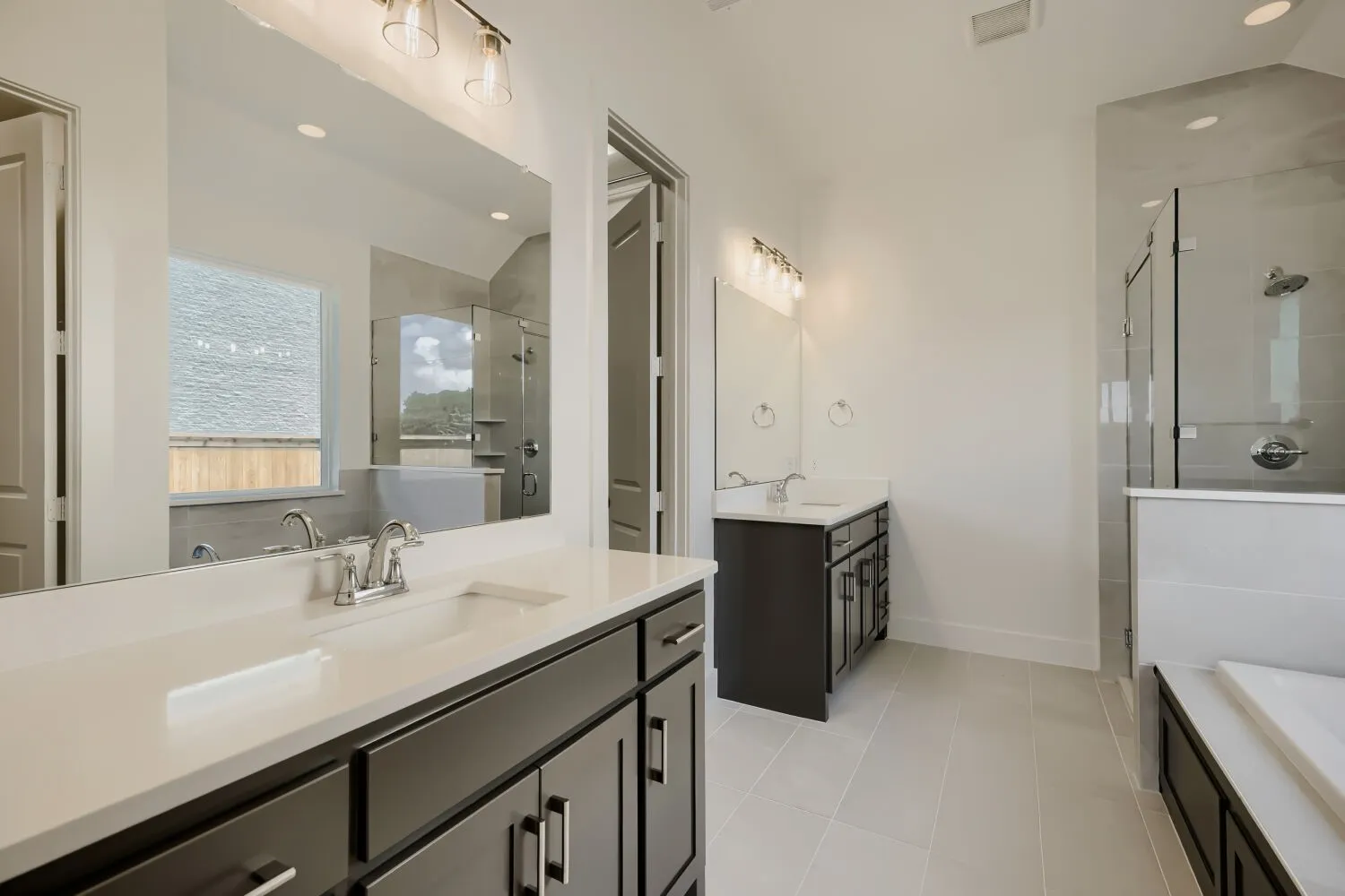 Dual dark wood-finish vanities with light countertops, chrome fixtures, a frameless glass shower enclosure, a built-in tub, and light tile flooring
