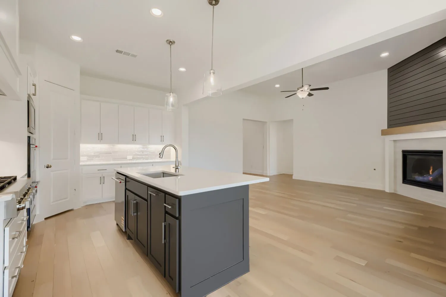 Open-concept kitchen and living area featuring light wood-finish flooring, a white kitchen with a dark gray island, a stainless steel range and dishwasher, an integrated fireplace with a shiplap accent wall, and recessed lighting