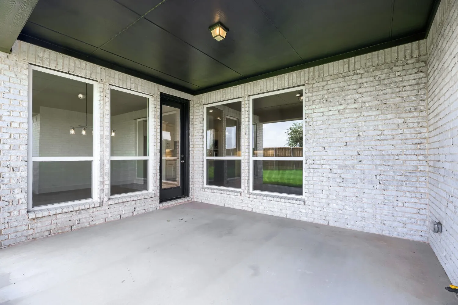 Covered outdoor patio featuring light-colored brick exterior, large windows, a full-light entry door, and a dark soffit with a flush-mount light fixture