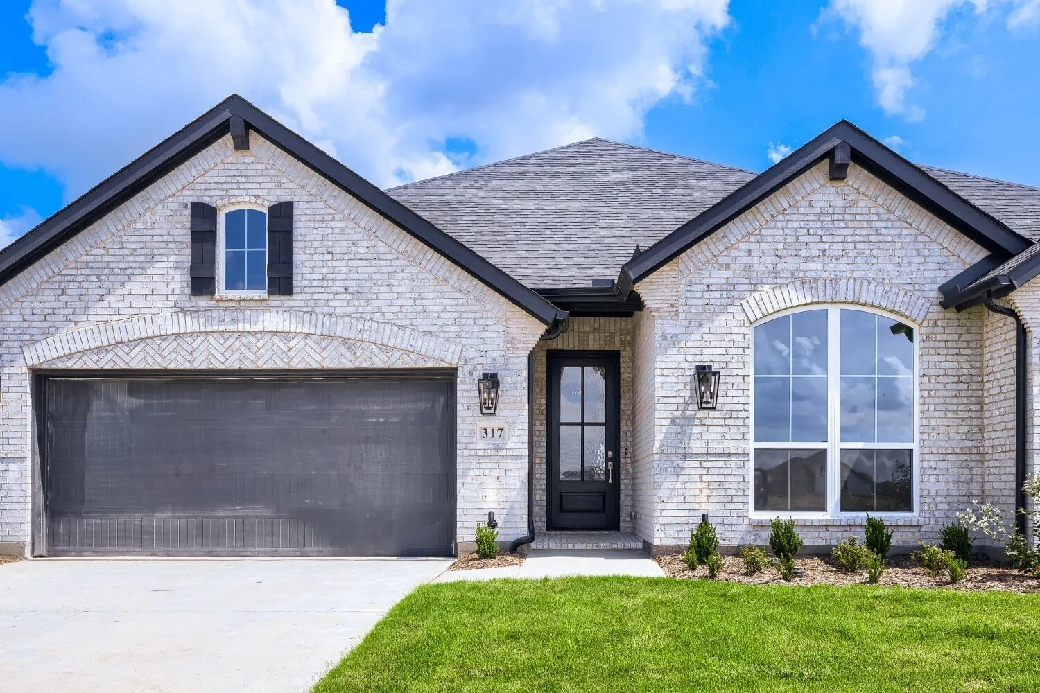 White brick exterior featuring a dark gray garage door, arched entry window, dark wood front door, and black exterior lighting fixtures