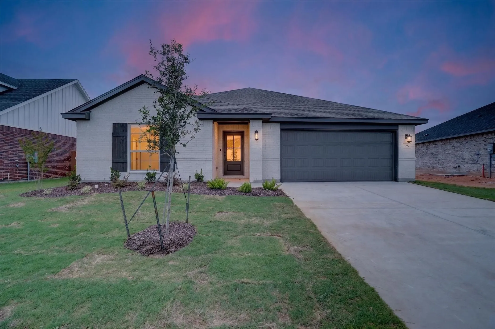 White brick facade with dark shutters, an attached garage, a paved driveway, and established landscaping