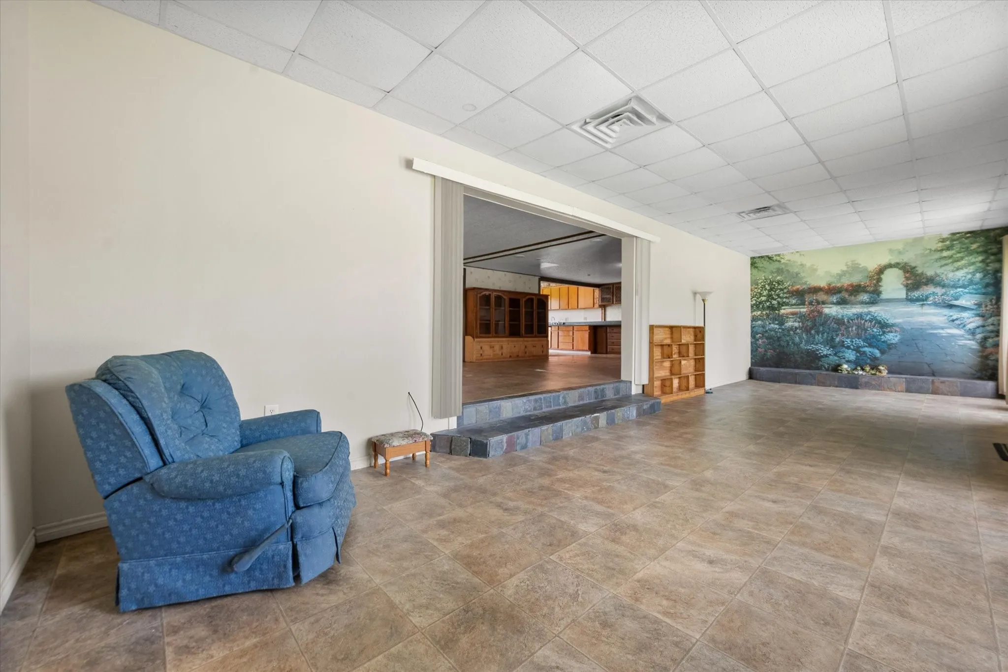 Spacious room featuring neutral-toned tile flooring, a drop ceiling, and a large wall mural depicting a garden scene
