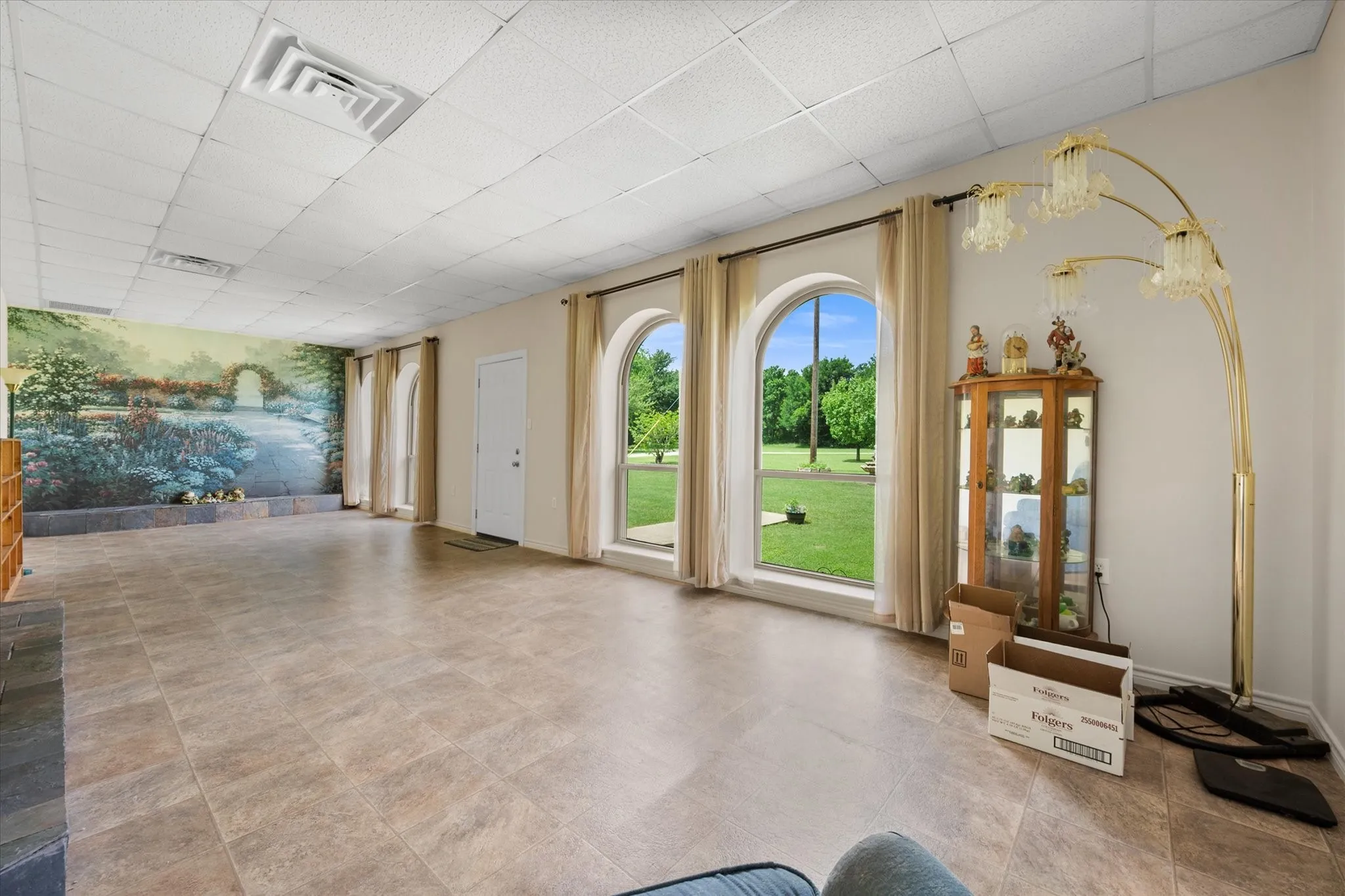 Spacious room featuring three large arched windows, tile flooring, a detailed wall mural, and a drop-tile ceiling
