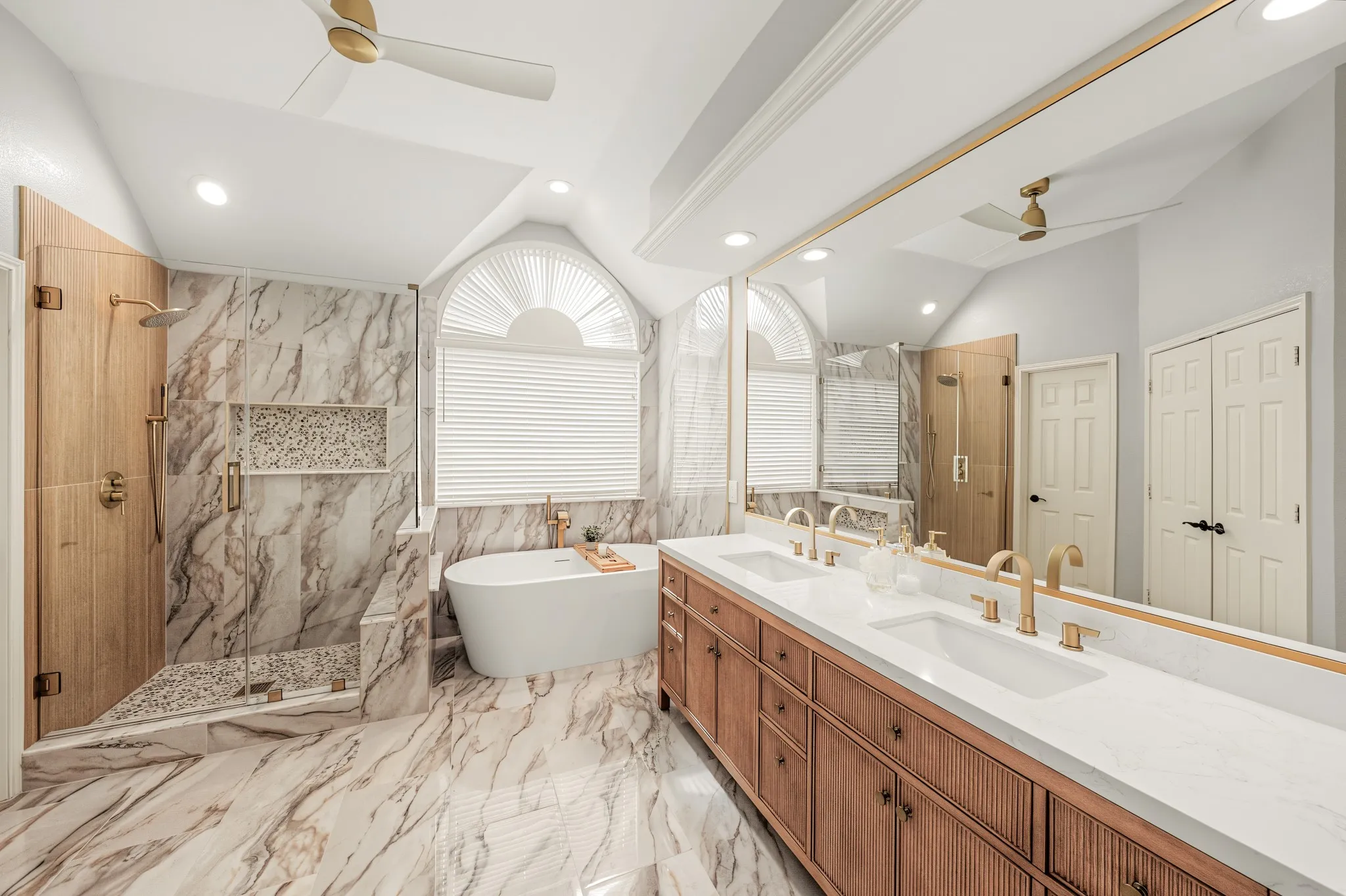 Spacious bathroom featuring a freestanding soaking tub, a walk-in shower with glass enclosure, a dual vanity with undermount sinks and white countertops, marble-look tile flooring, and a large arched window with blinds