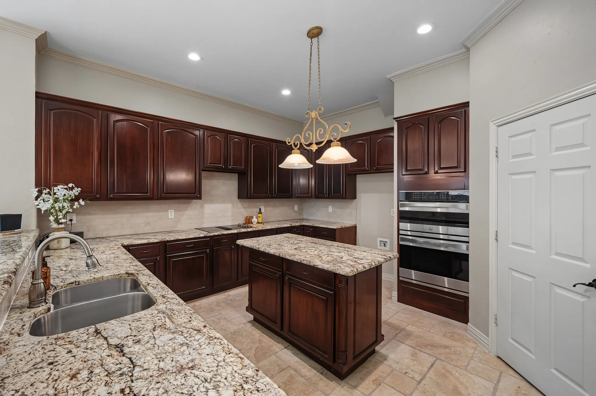 Kitchen featuring rich wood cabinetry, stone countertops, a double basin stainless steel sink, a built-in double oven, and a central island with additional storage