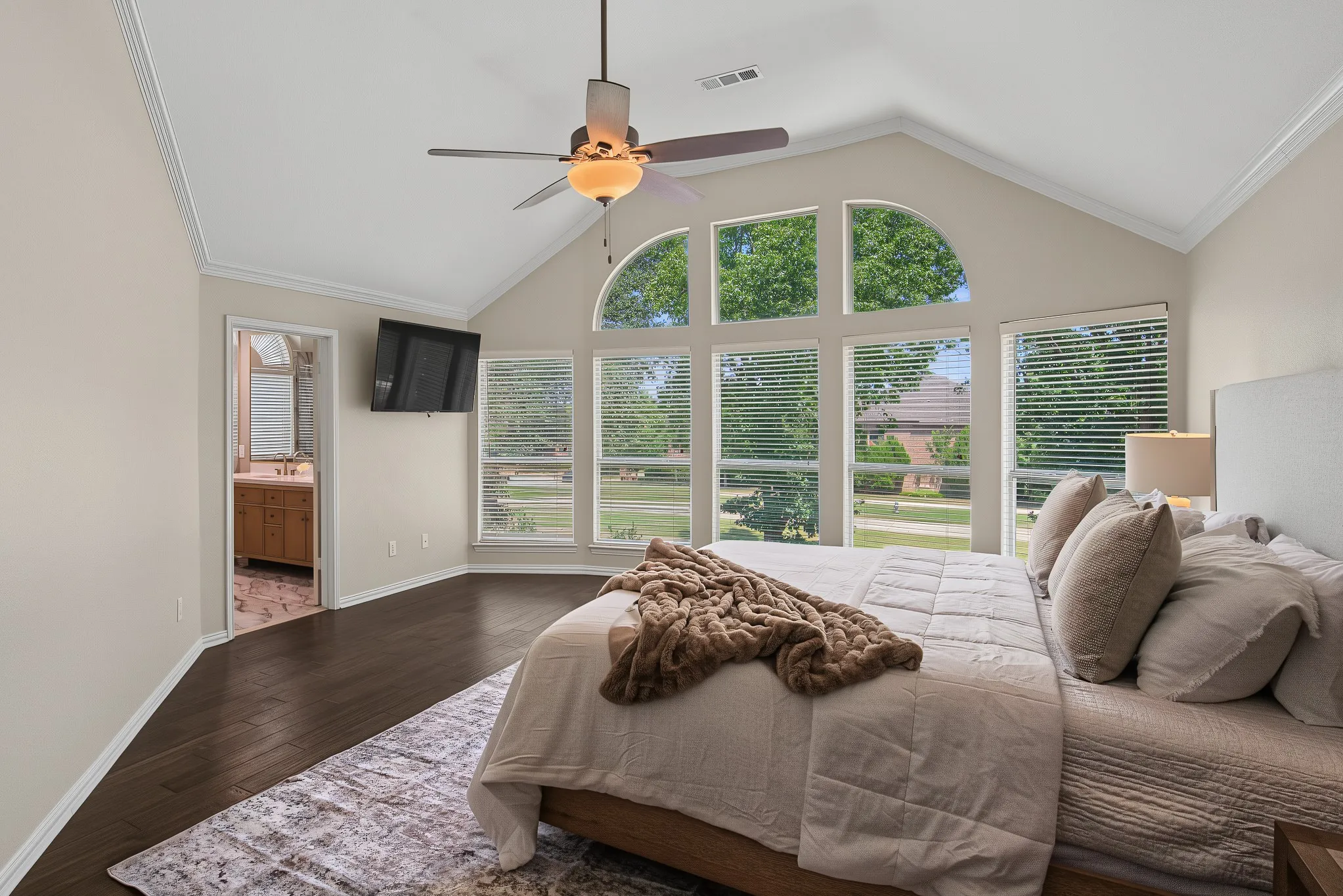 Vaulted ceiling with crown molding, dark wood-finish flooring, large arched and rectangular windows, ceiling fan, and recessed wall-mounted television