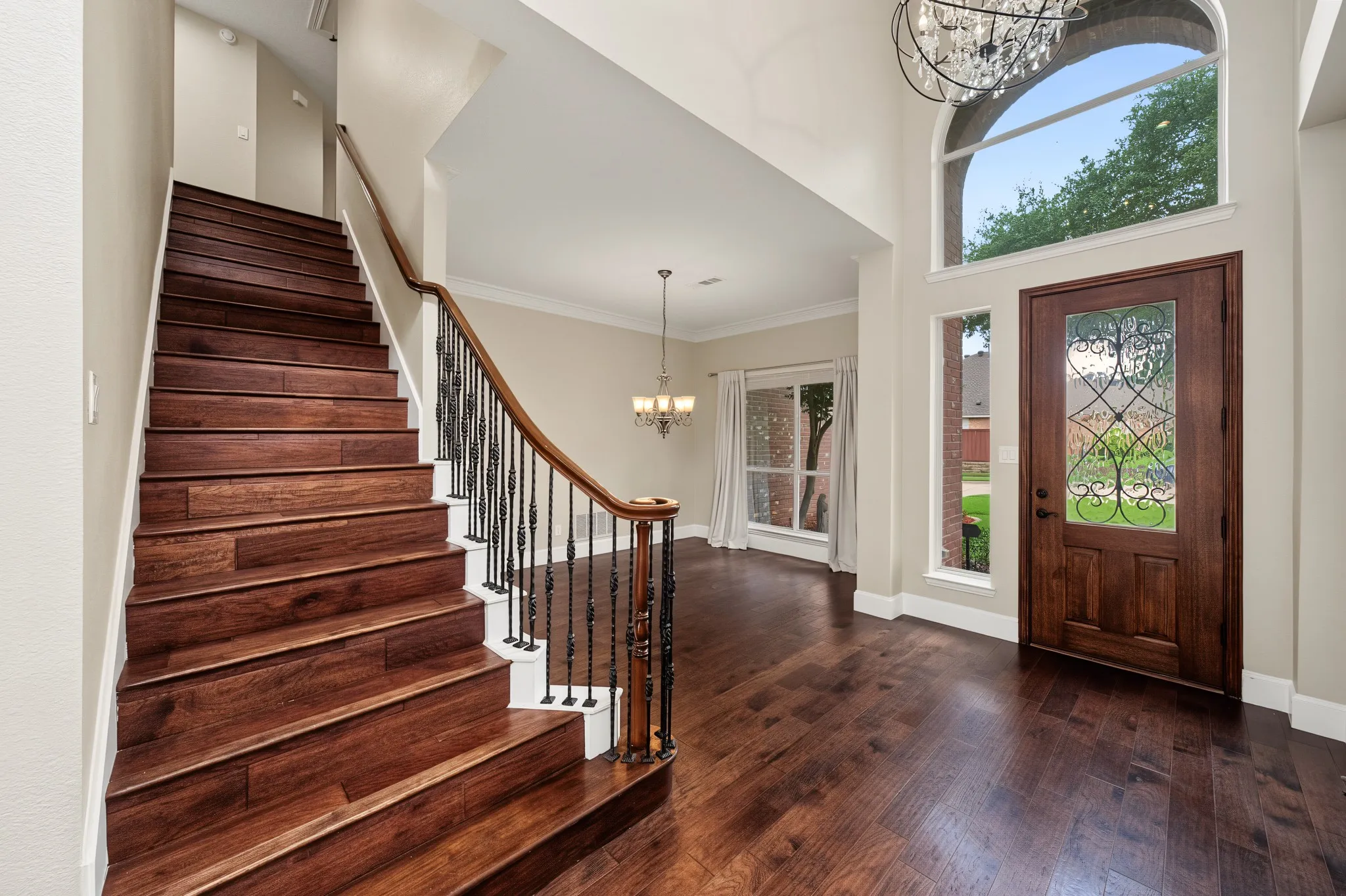 Grand entry featuring rich wood-finish flooring, a substantial wood staircase with wrought iron balusters, a large arch-top window, and a chandelier