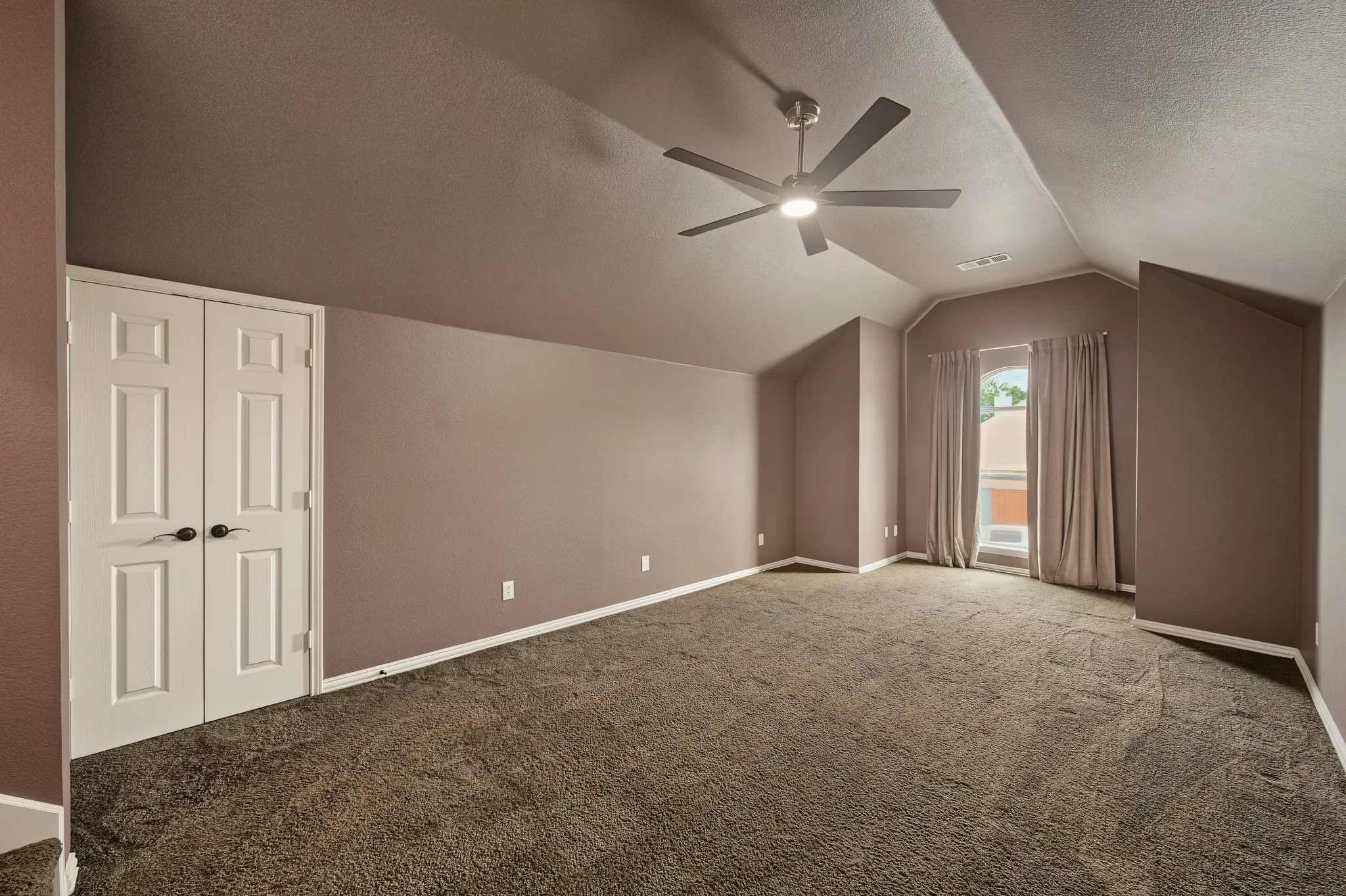 Spacious room featuring vaulted ceilings, a contemporary ceiling fan with integrated lighting, window with drapes, and plush carpet flooring