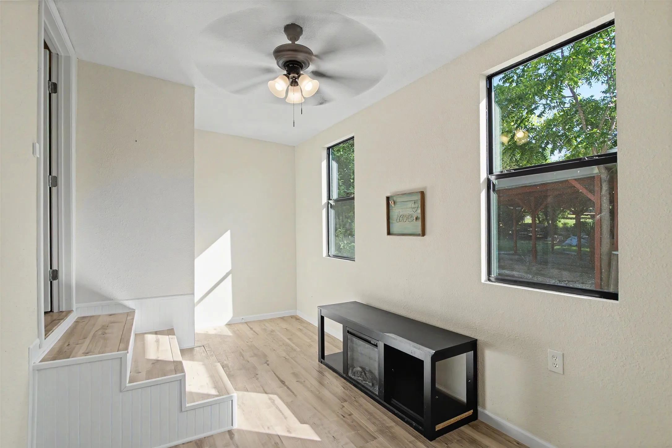 Room featuring wood-finish flooring, two large windows, a ceiling fan with integrated lighting, and a built-in step structure with wainscoting