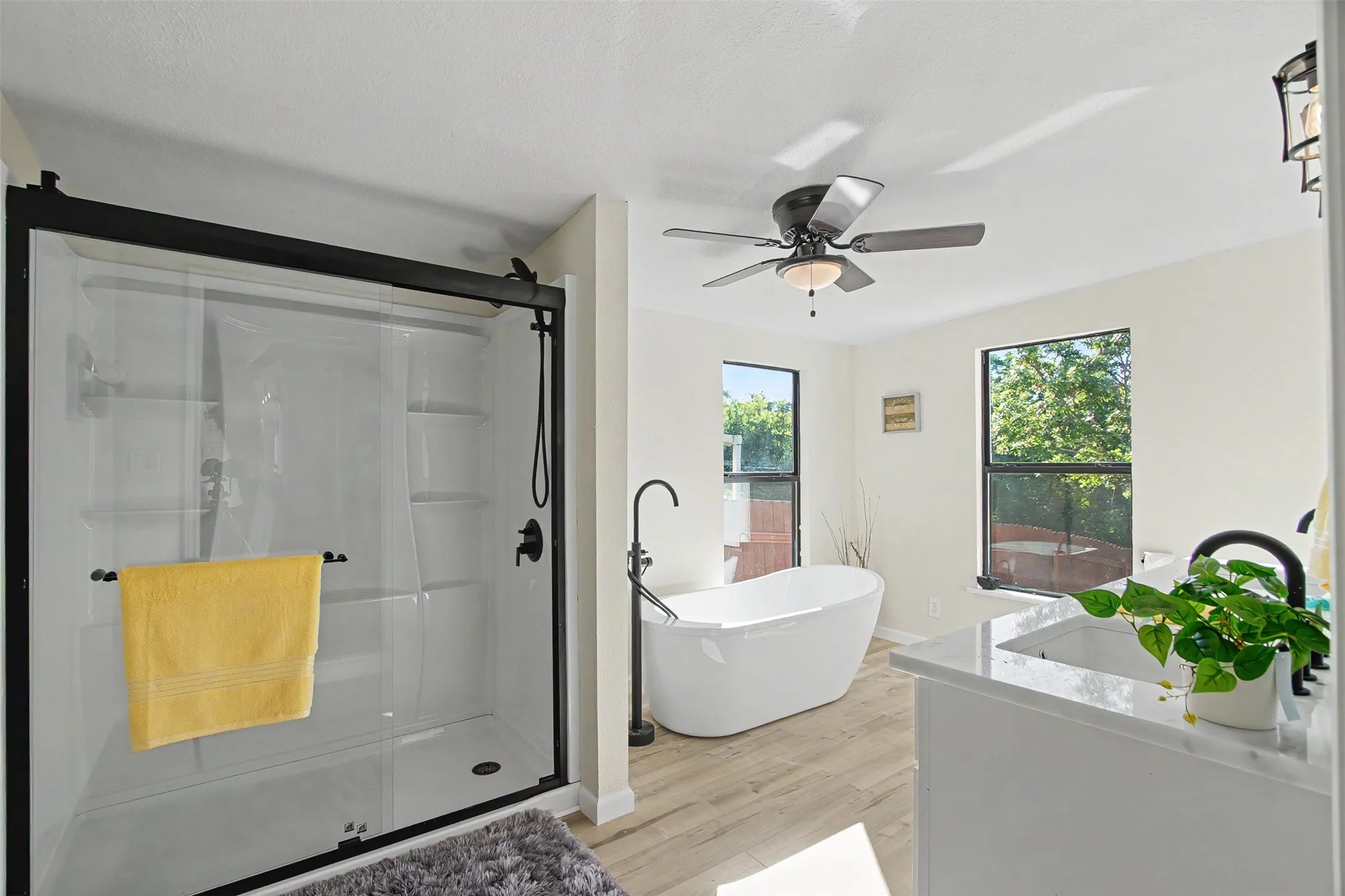 White bathroom featuring a glass-enclosed shower with black fixtures, a freestanding soaking tub, wood-finish flooring, and a ceiling fan