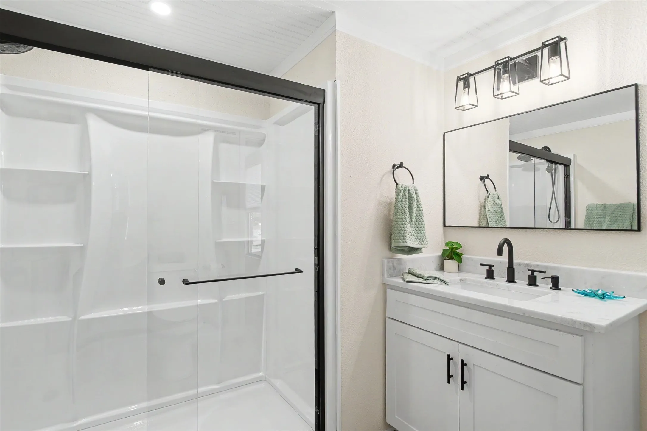 Walk-in shower with clear glass enclosure and integrated shelving, accompanied by a vanity with a rectangular undermount sink, stone countertop, matte black faucet, and coordinating cabinet hardware