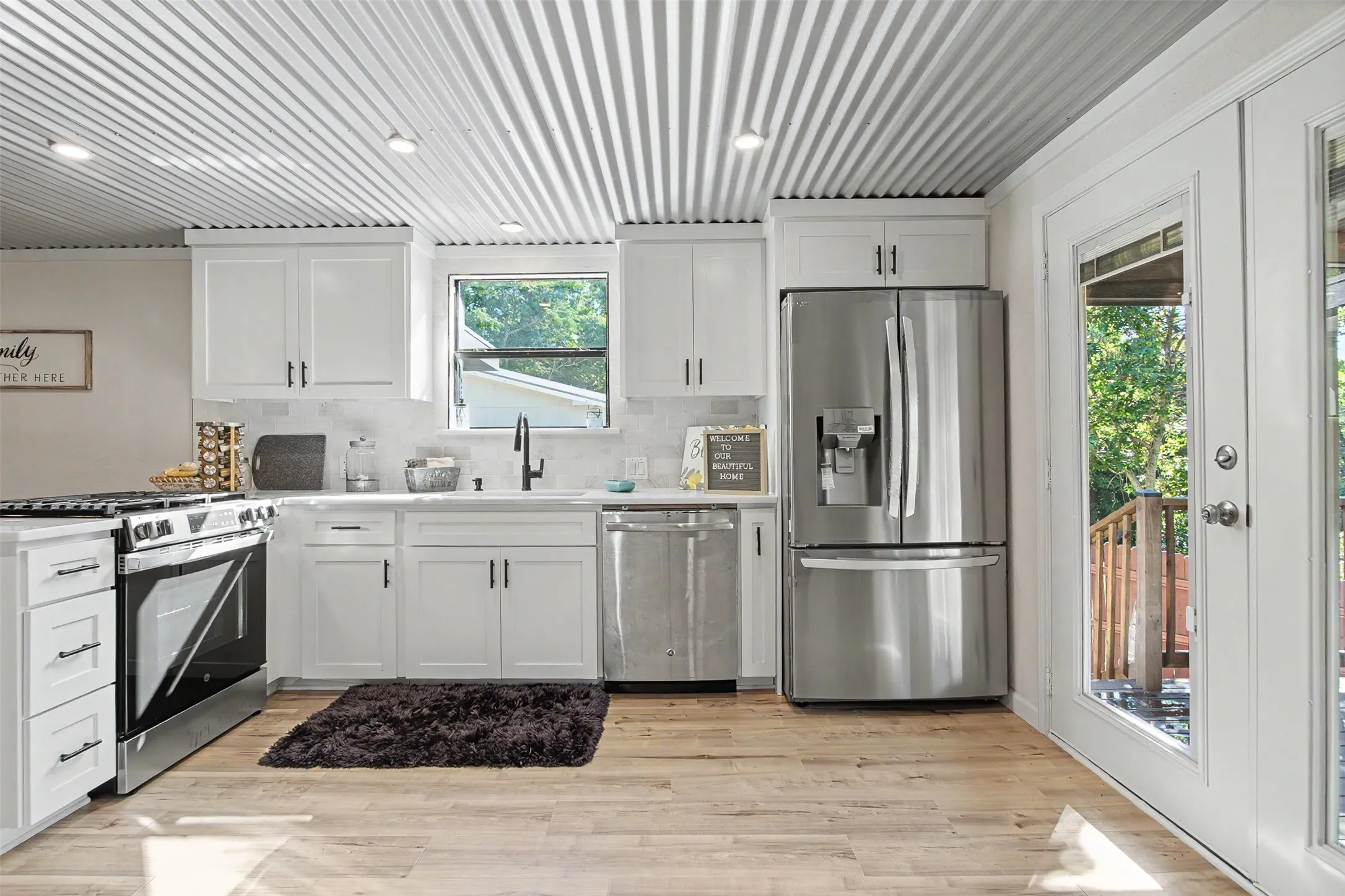 Kitchen featuring a corrugated metal ceiling, wood-finish flooring, white cabinetry with dark hardware, stainless steel appliances, and a glass-paneled exterior door