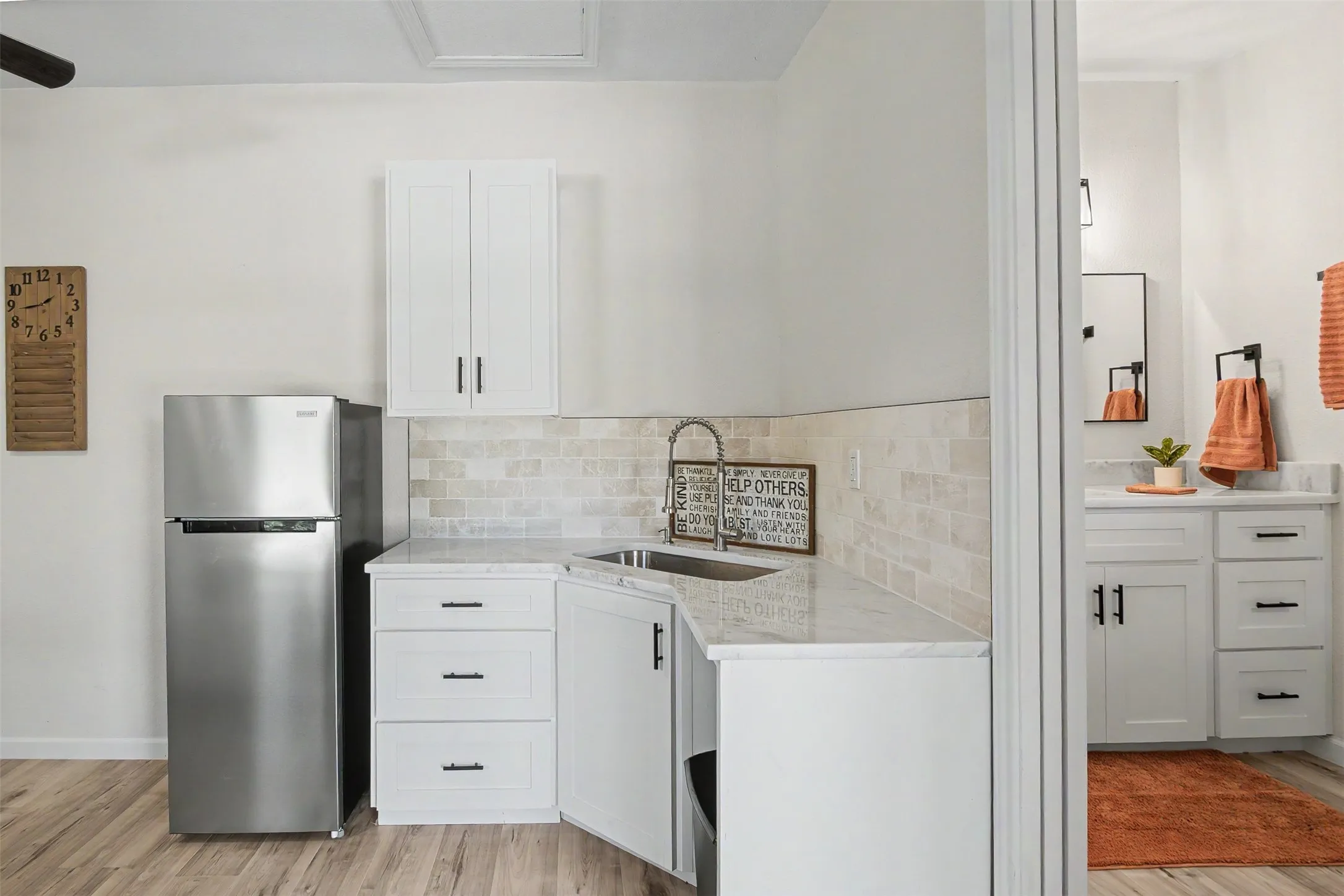Compact kitchen area featuring white shaker-style cabinetry with dark hardware, a stainless steel sink with a gooseneck faucet, light-toned tiled backsplash, and wood-finish flooring